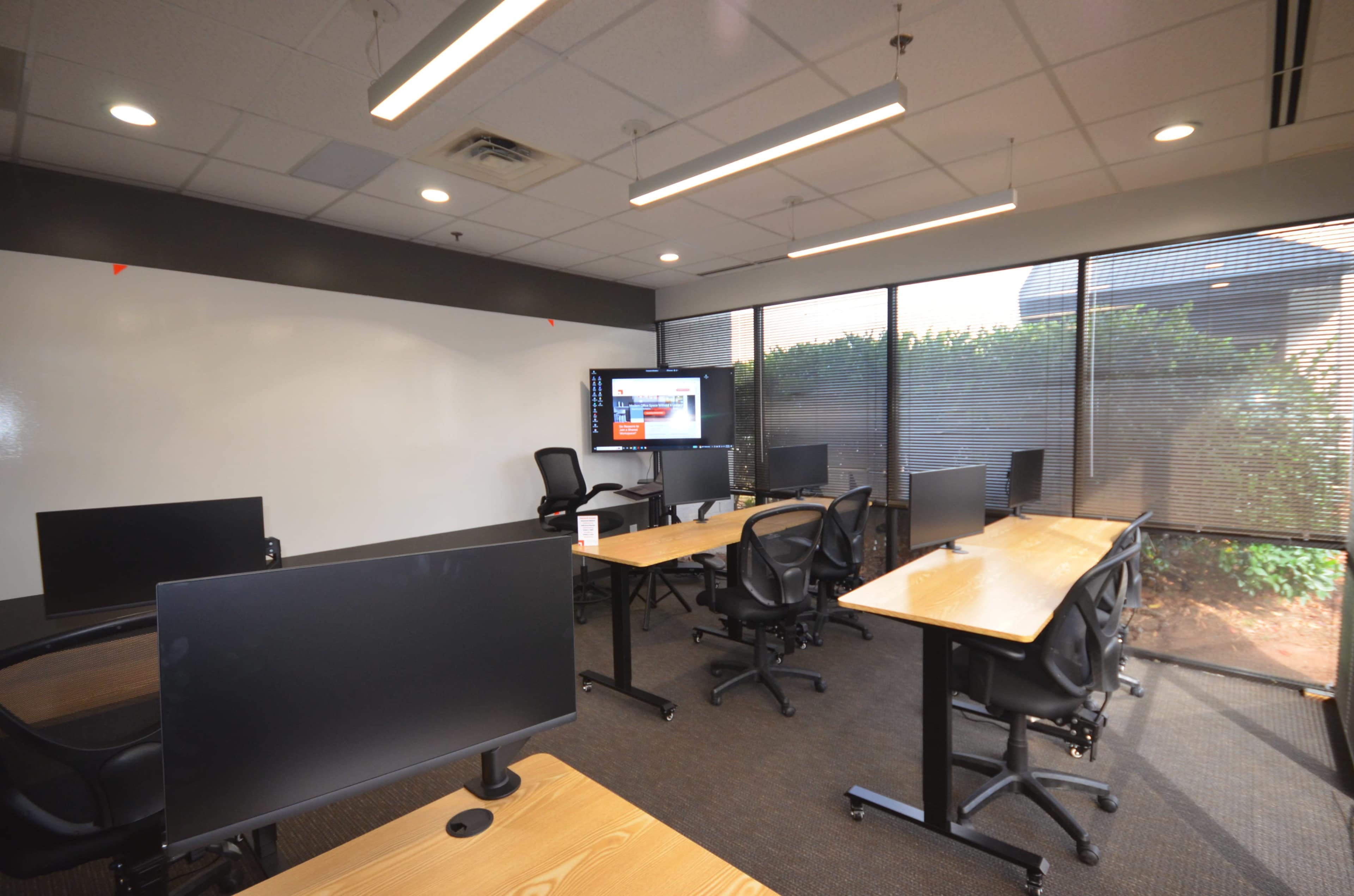 A modern classroom features multiple desks with computers, a large screen, and windows providing natural light.