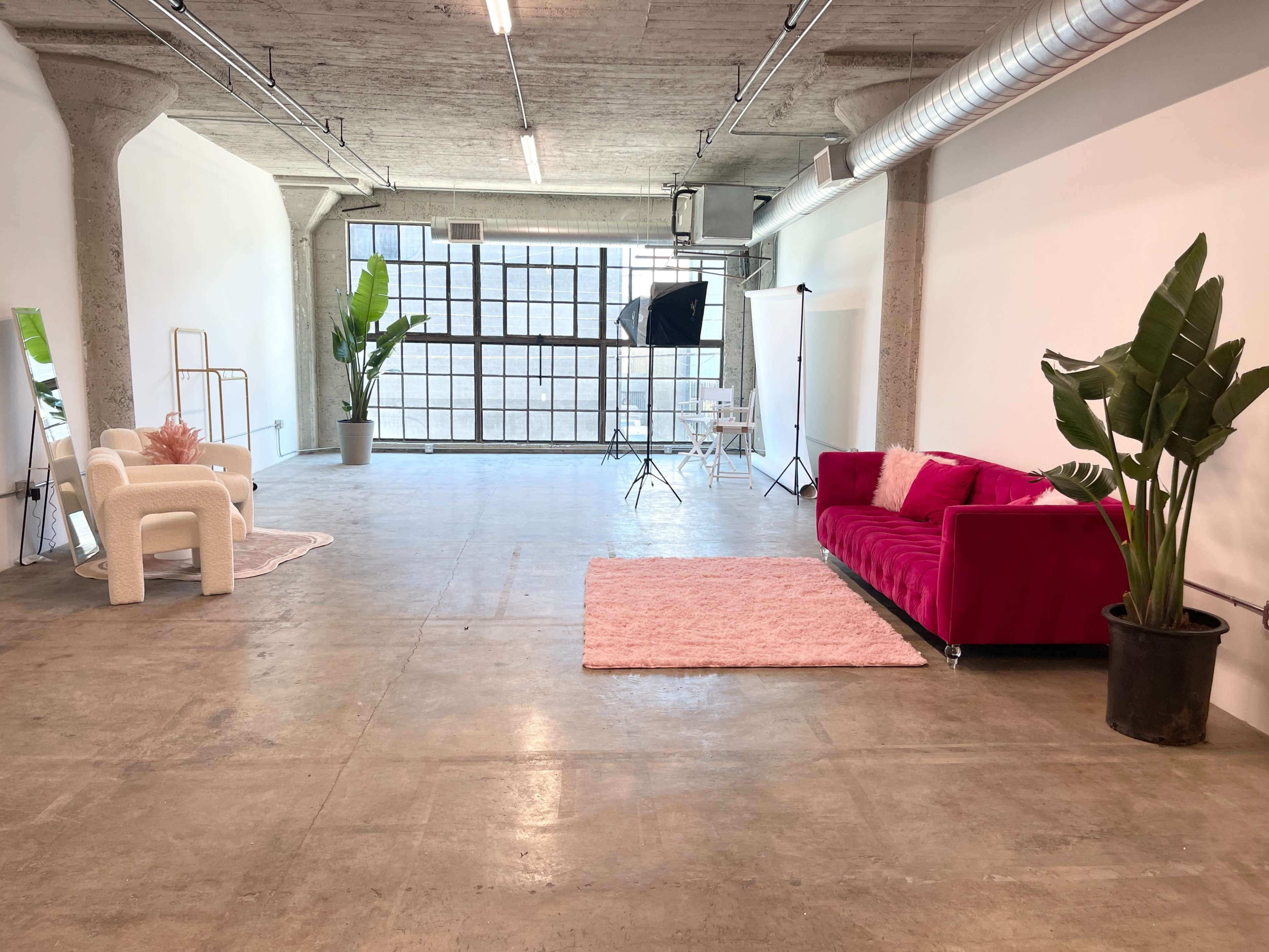 The image depicts a spacious industrial-styled room with concrete floors, large windows, a fuchsia couch, a white chair, and potted plants, along with photography equipment in the background.