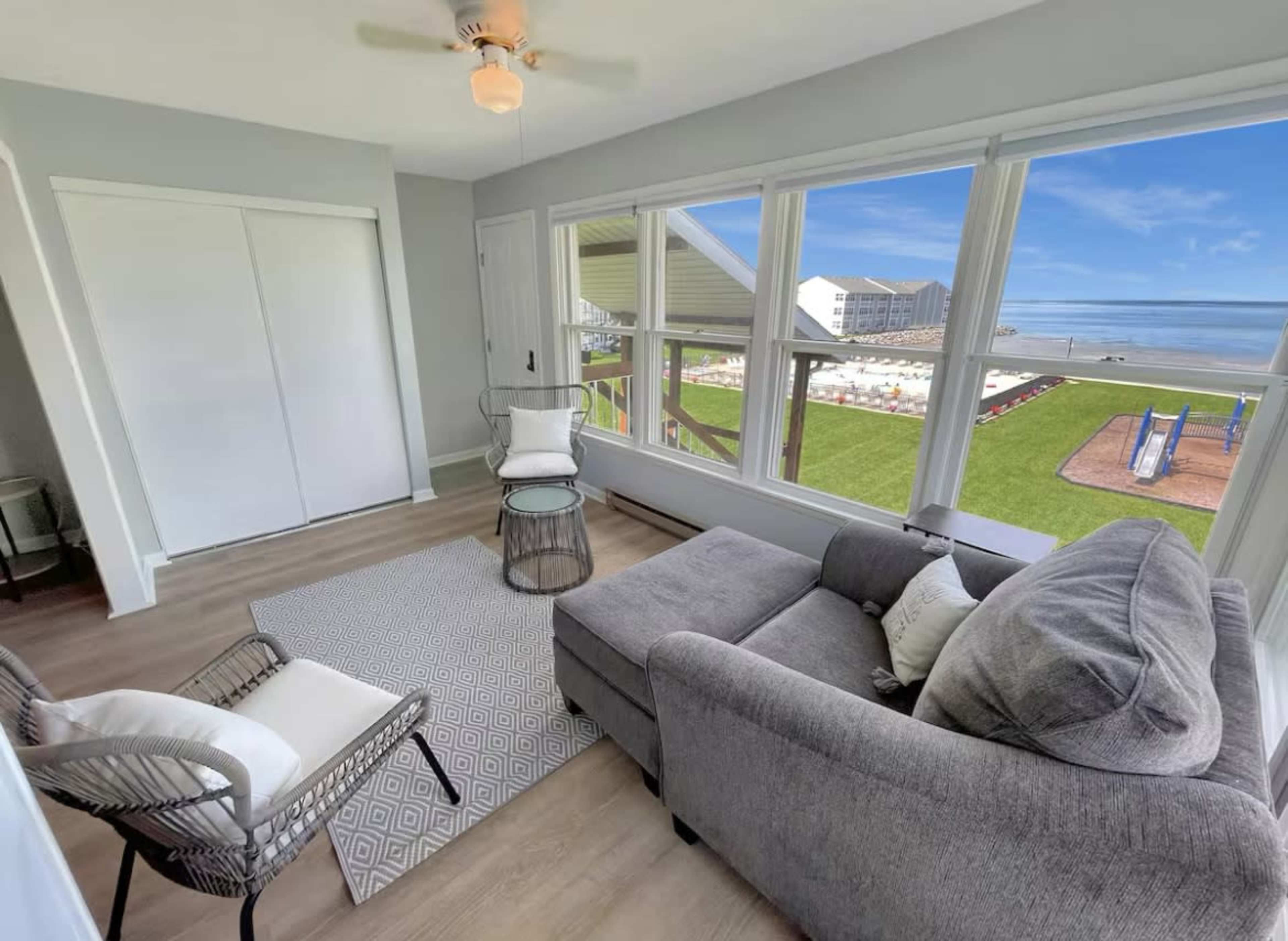 A cozy seating area with a gray sofa and a small table, featuring large windows that overlook a grassy outdoor space and beach.