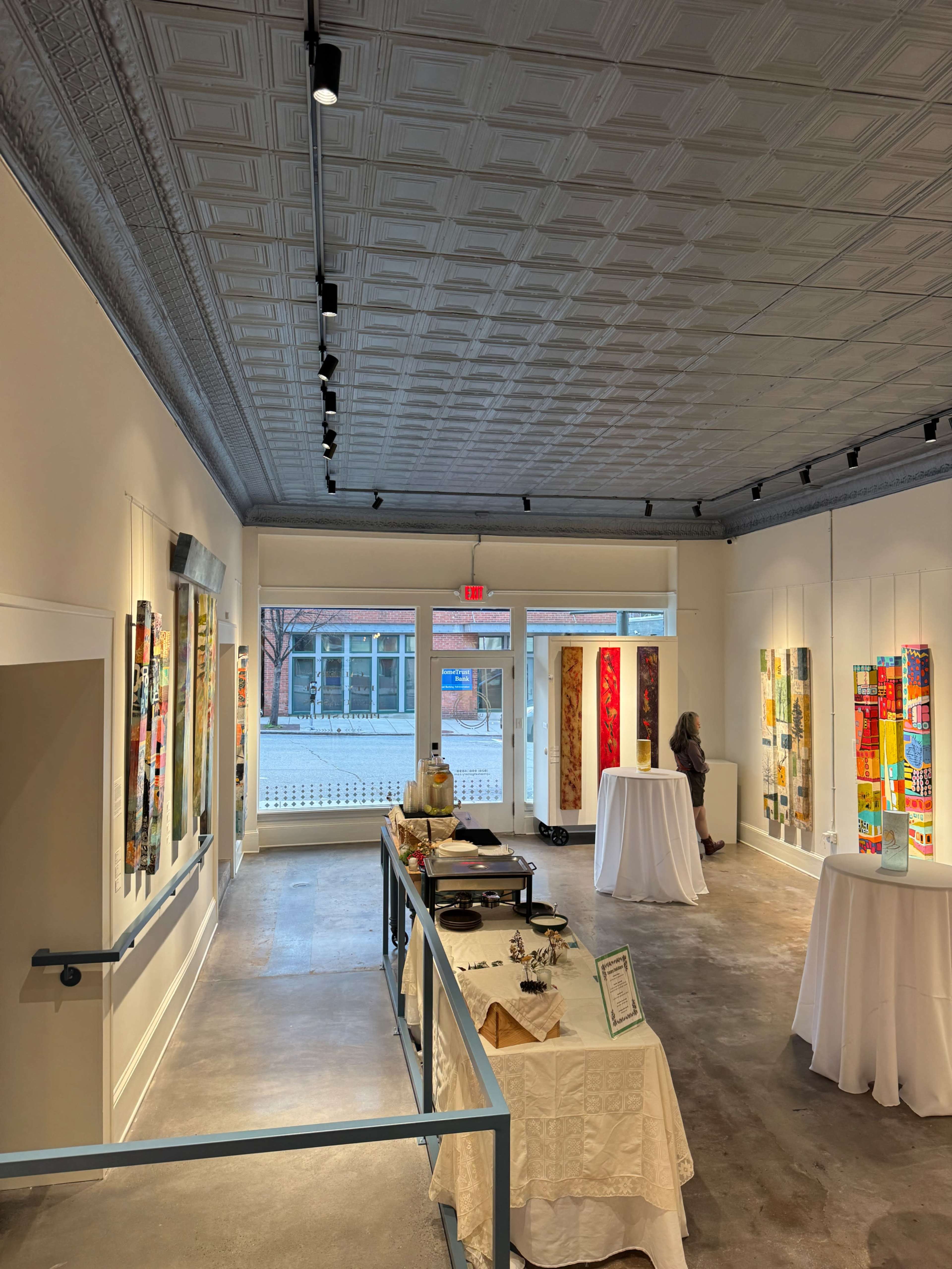 The image shows the interior of an art gallery featuring colorful artworks displayed on the walls, with tables set up for an event and people moving through the space.