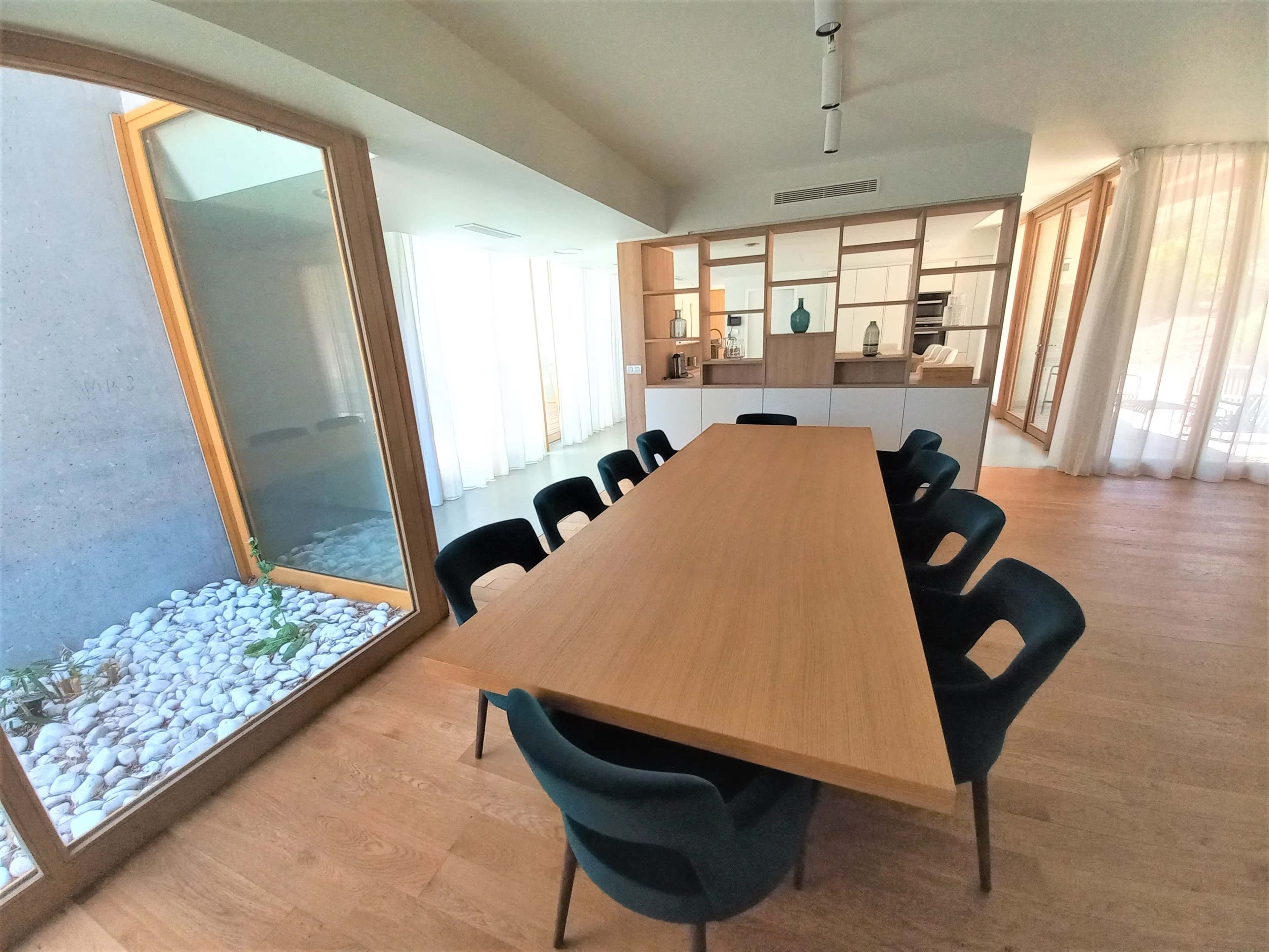 A spacious dining area features a long wooden table surrounded by black chairs, with large windows allowing natural light and a view of a pebble-filled garden.