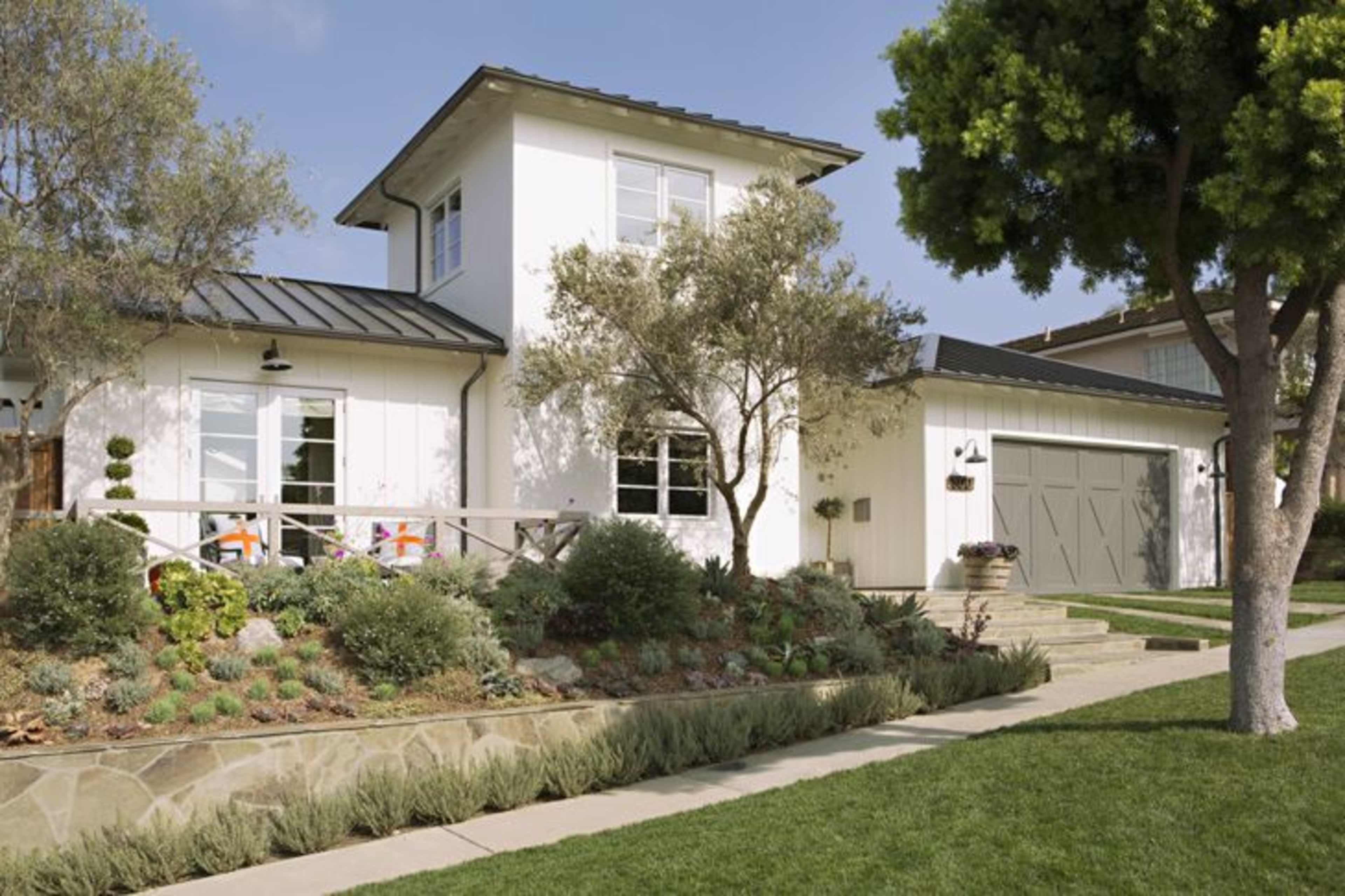 A two-story white house with a dark metal roof is surrounded by landscaped gardens and a driveway leading to a garage.