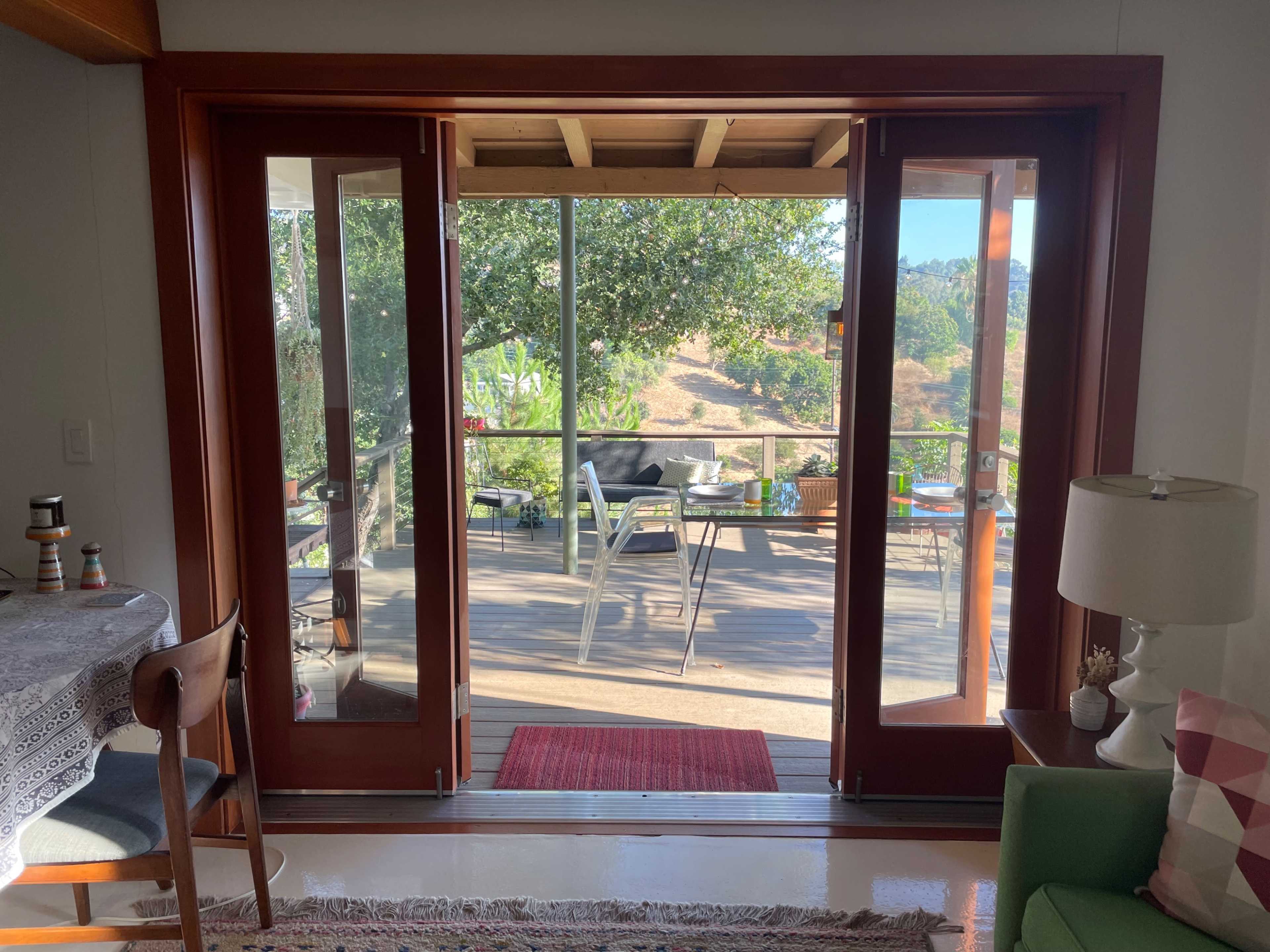 A set of double glass doors opens to a wooden deck with outdoor furniture and greenery.
