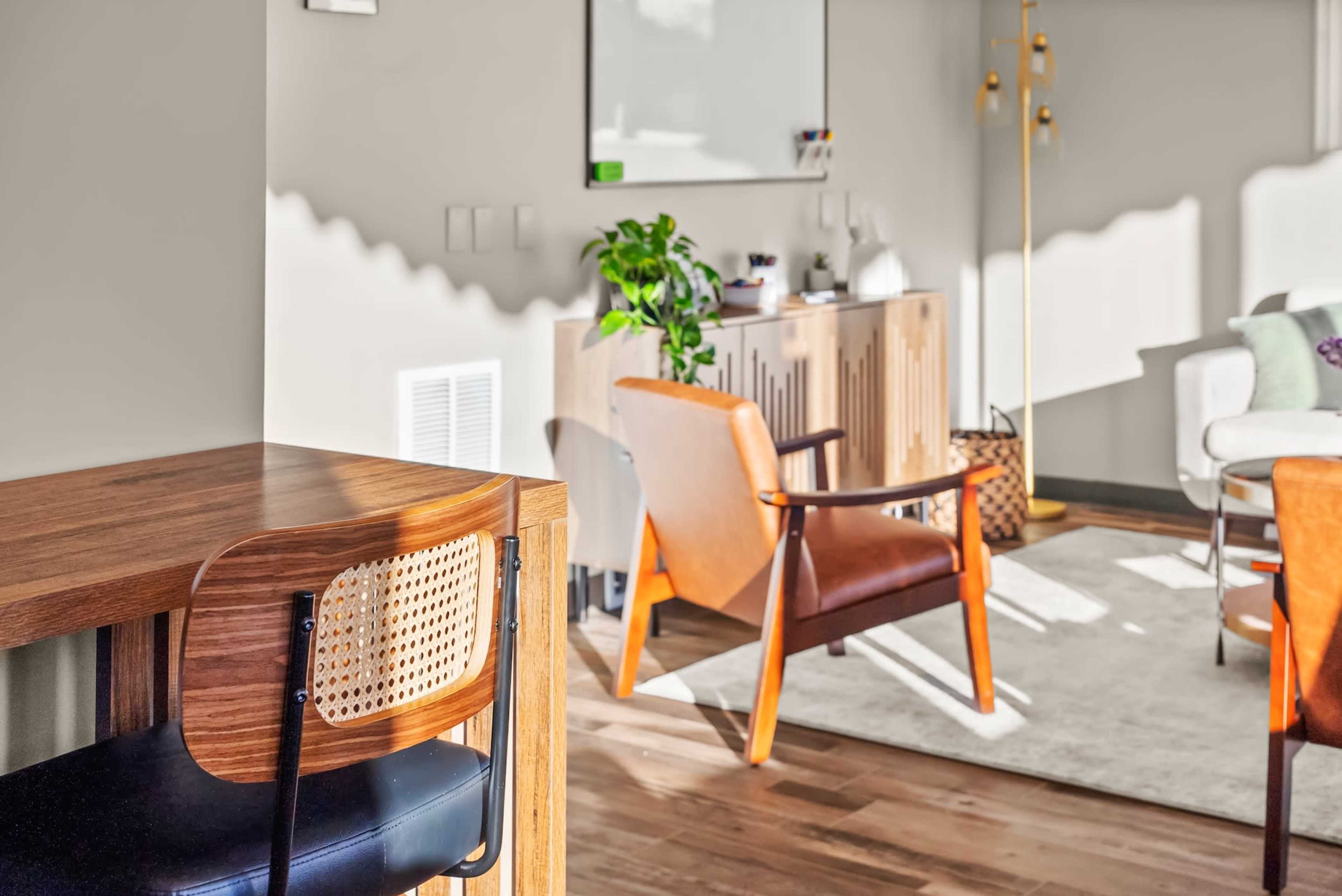 The image shows a bright, modern interior with a wooden table, leather chairs, a light rug, and a green plant in a well-lit living space.