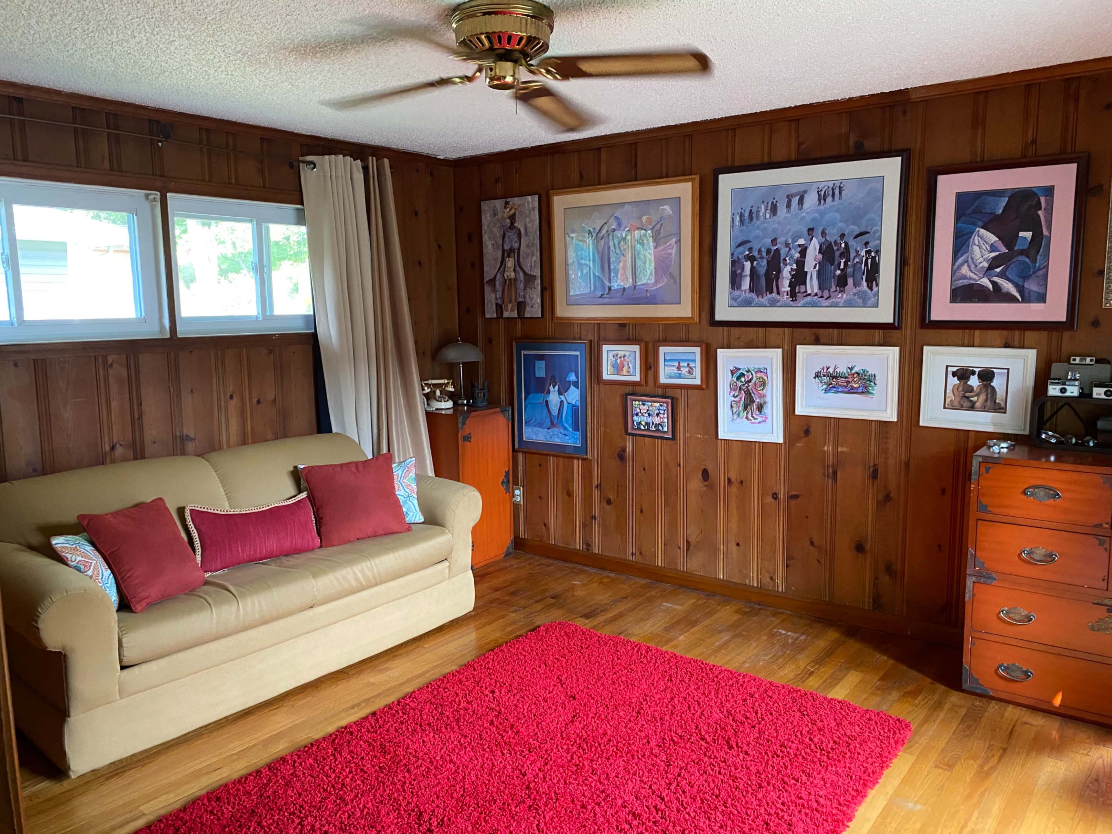 A cozy interior room featuring a light-colored sofa with red pillows, a red area rug, and a wall adorned with various framed artworks and photographs.
