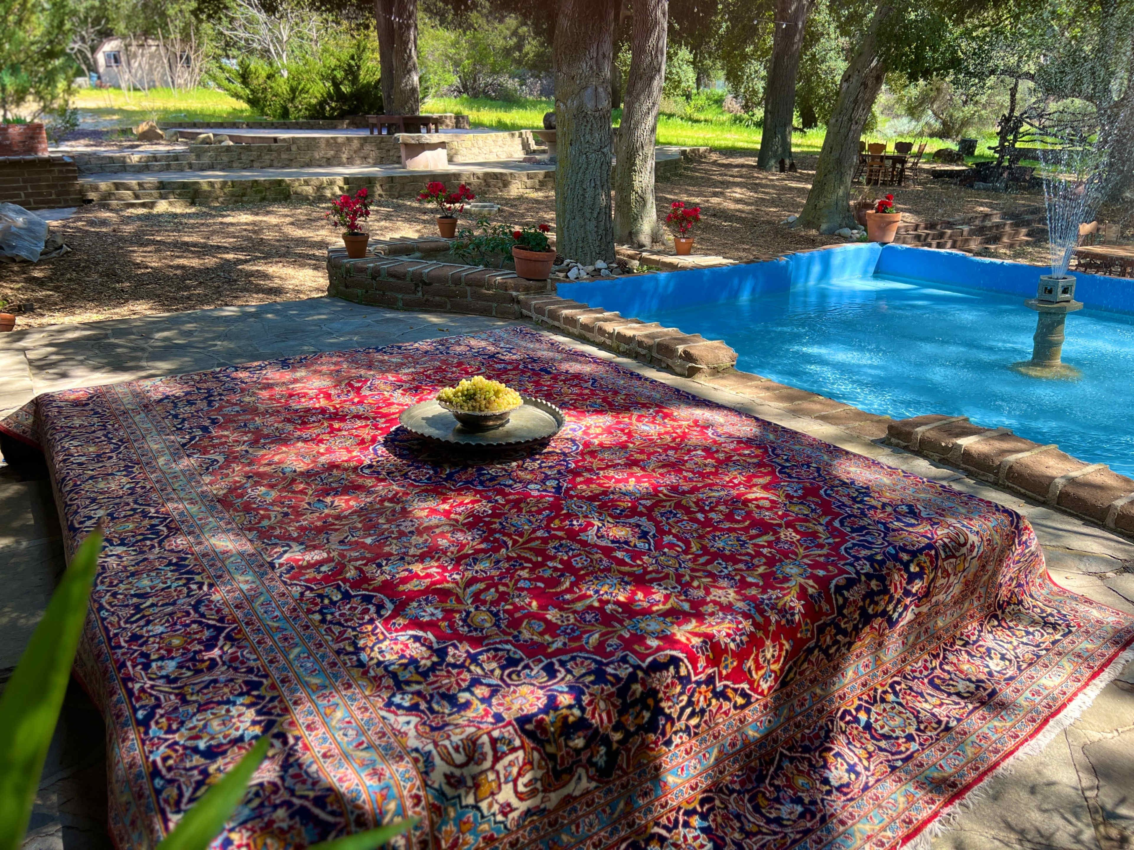 A vibrant rug is spread out on a stone patio, with a bowl of fruit placed in the center, surrounded by a swimming pool and lush greenery.