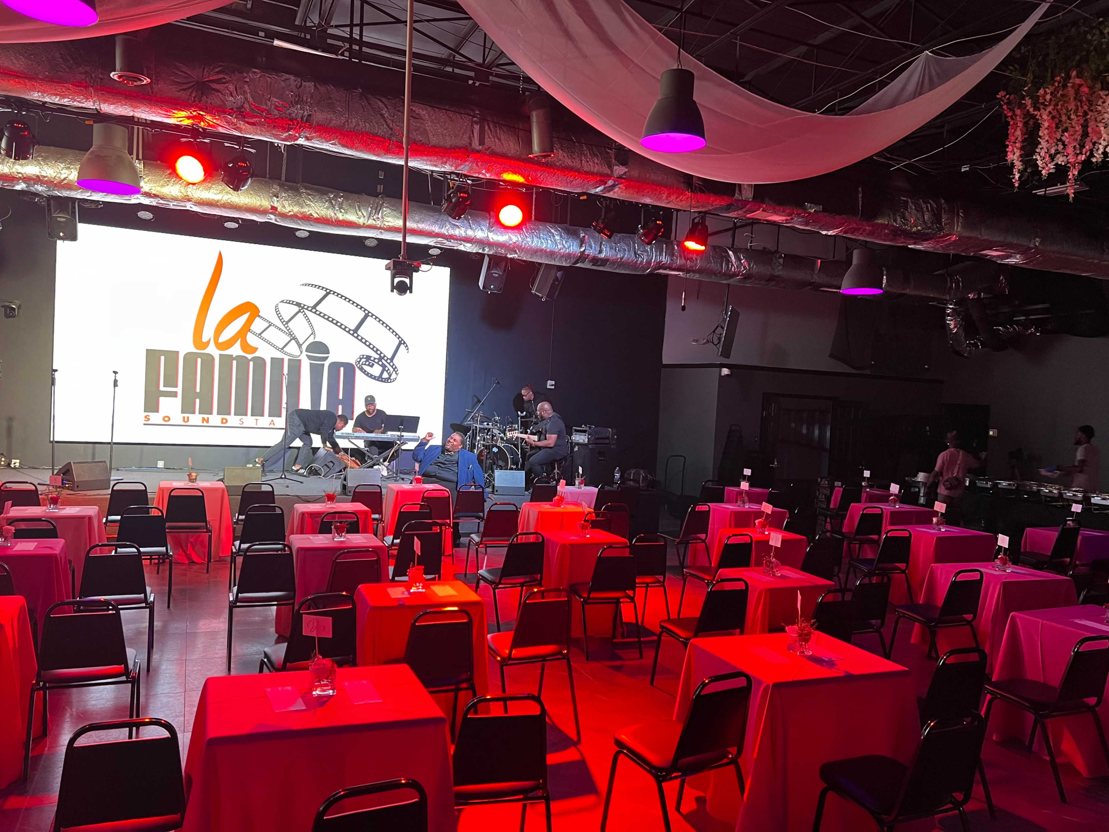 The image shows an empty event space set up for a concert, featuring several tables with pink tablecloths and a stage in the background displaying the logo "la familia."