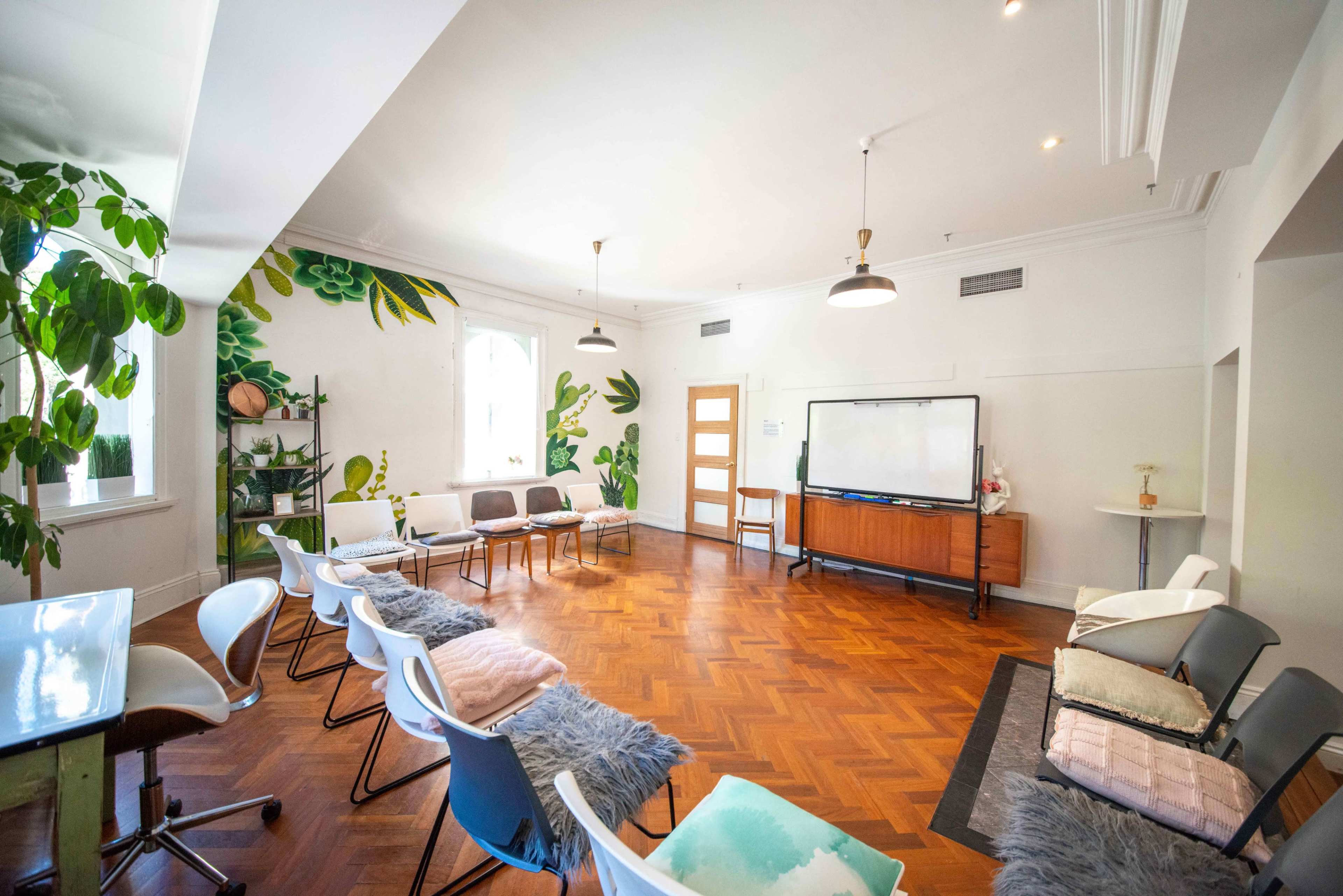 A spacious room with hardwood floors features a series of chairs arranged around a central area, a large whiteboard, and green plant murals on the walls.
