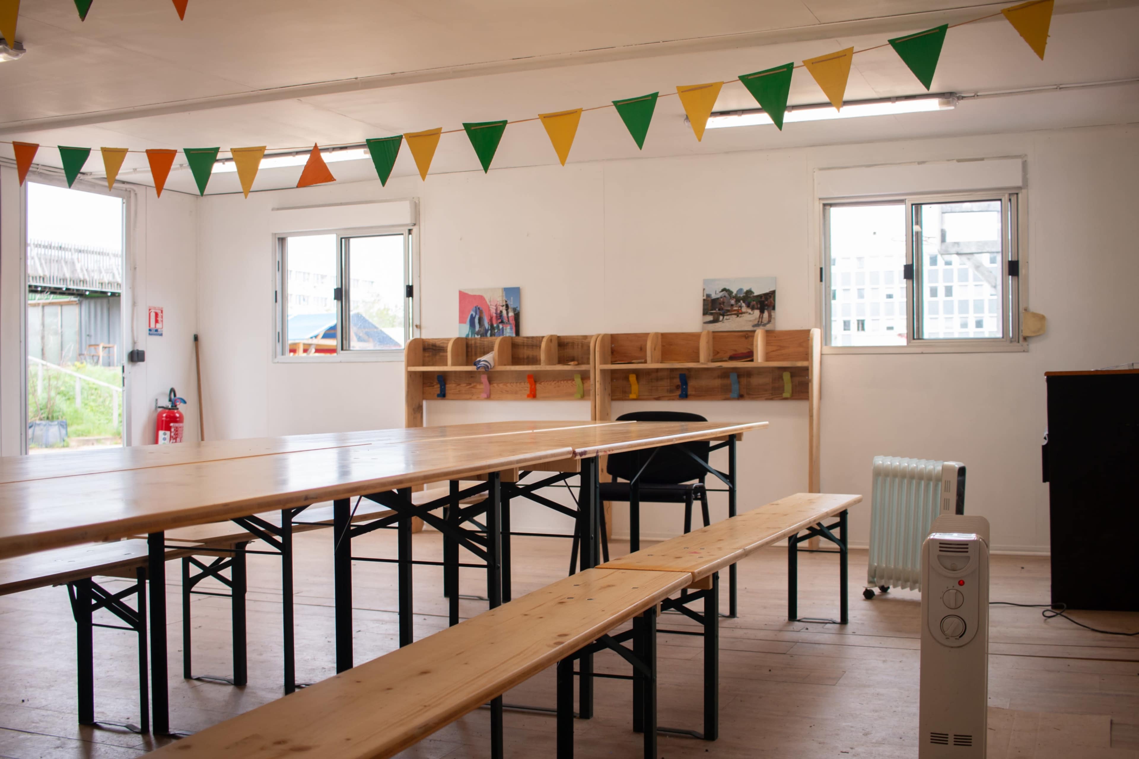 An indoor space with wooden tables and benches, decorated with colorful bunting and featuring windows and a shelf with pictures.