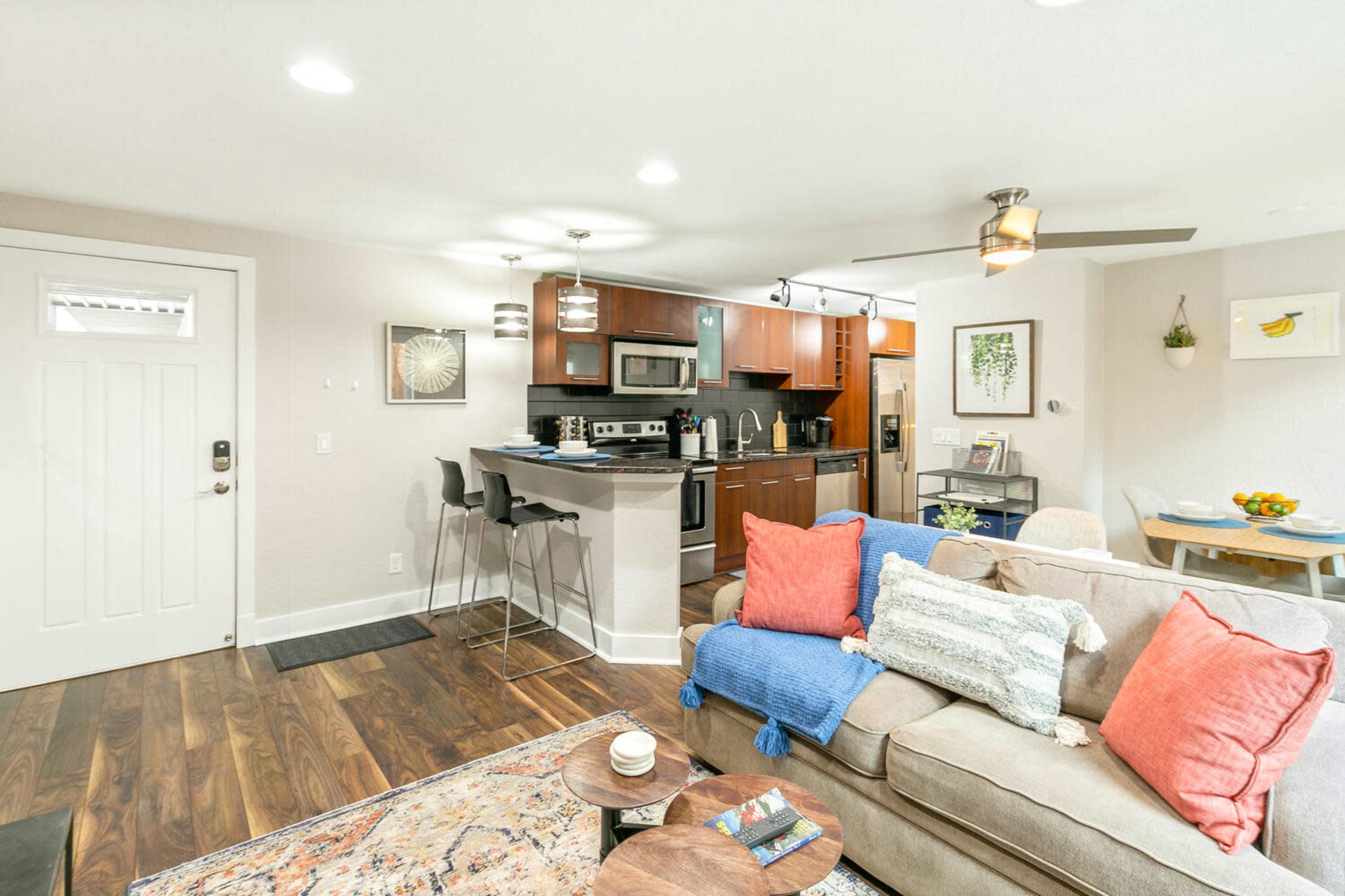 The image shows a modern living space that includes a kitchen area with dark wood cabinets, a cozy sofa with colorful cushions, and a dining table in a well-lit room.