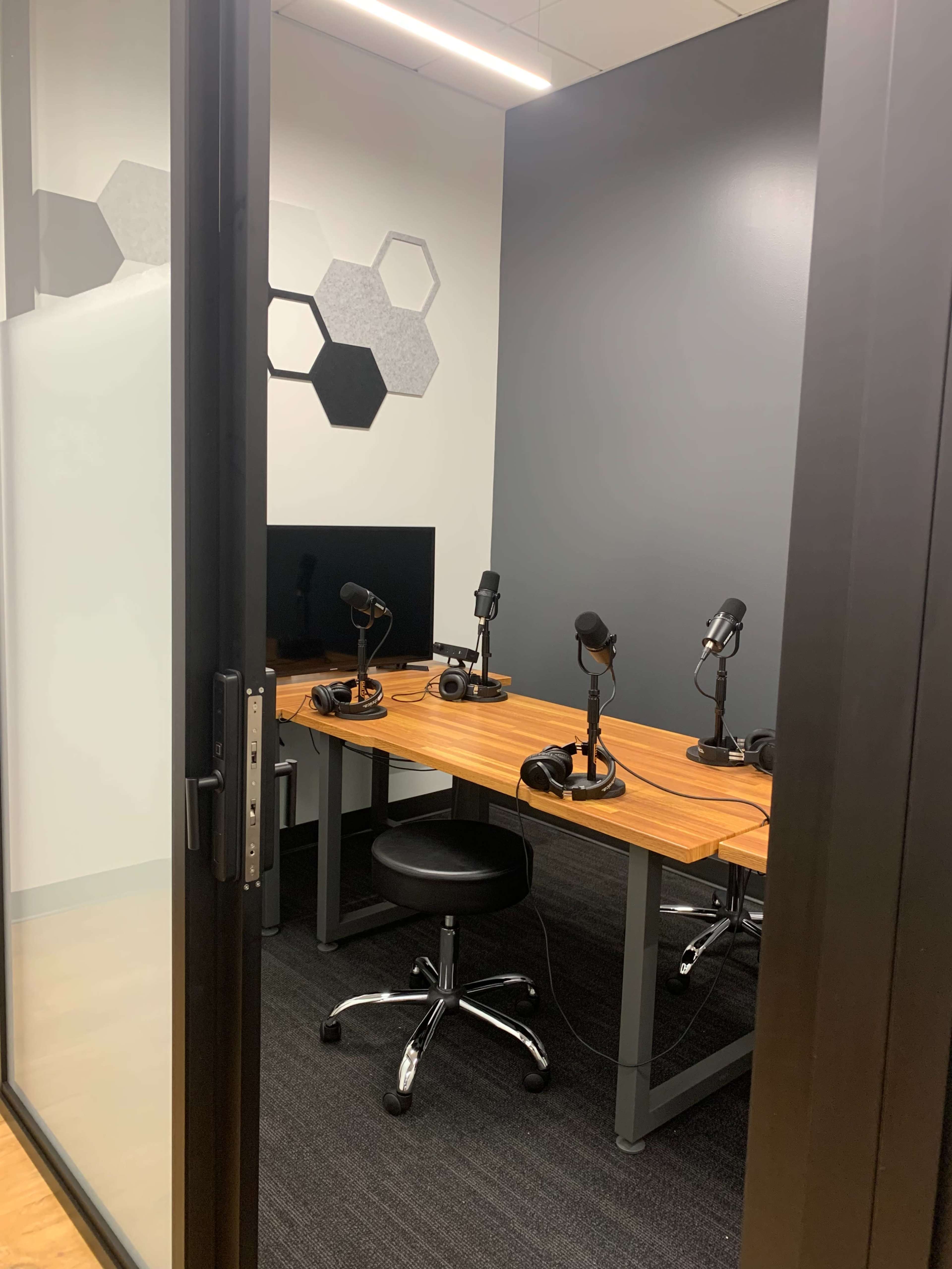 A small recording studio with a wooden table, multiple microphones, a monitor, and a black rolling chair, enclosed by glass doors.
