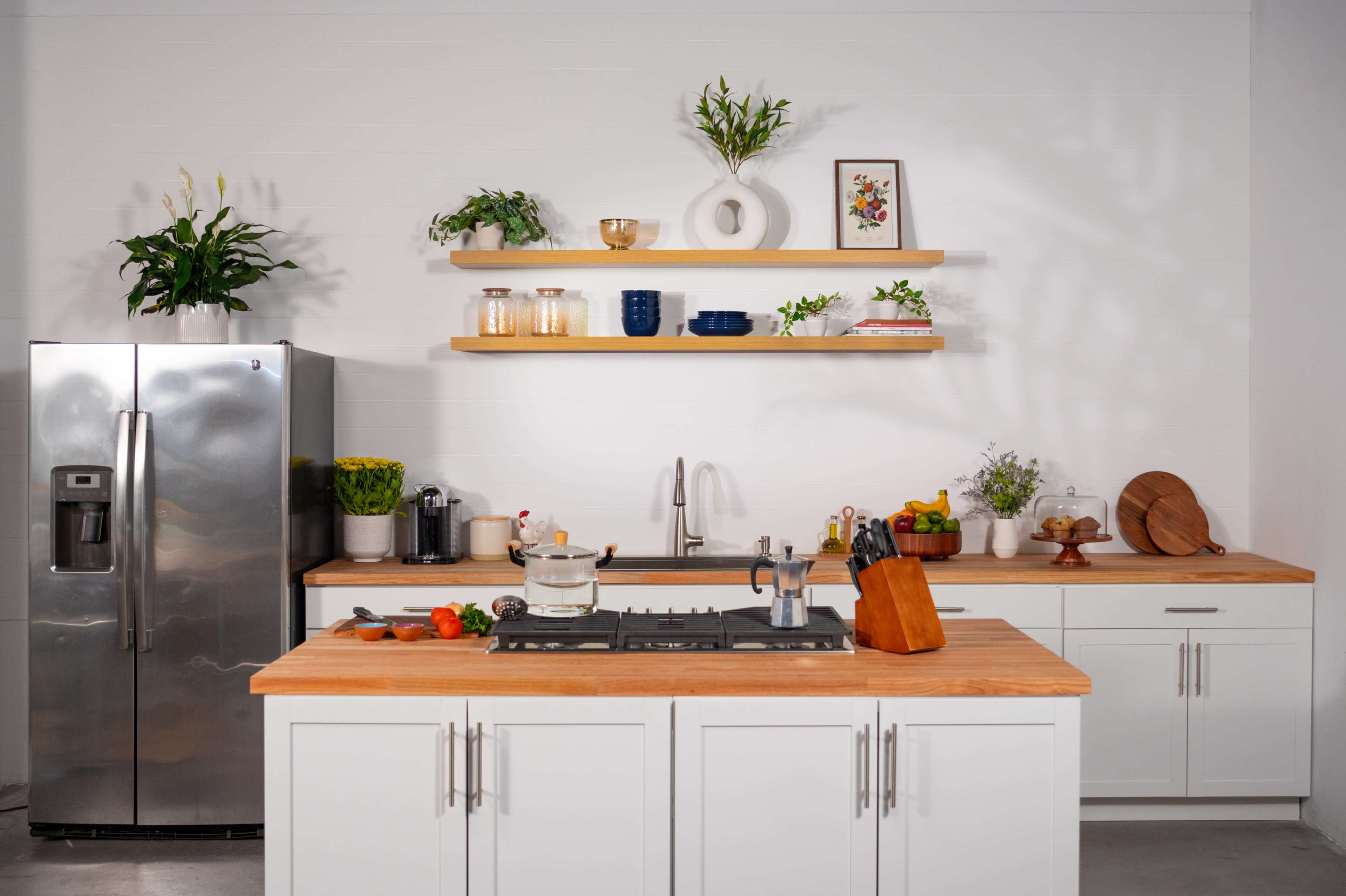 A modern kitchen features a stainless steel refrigerator, wooden countertops, and shelves displaying various kitchenware and decorative plants.
