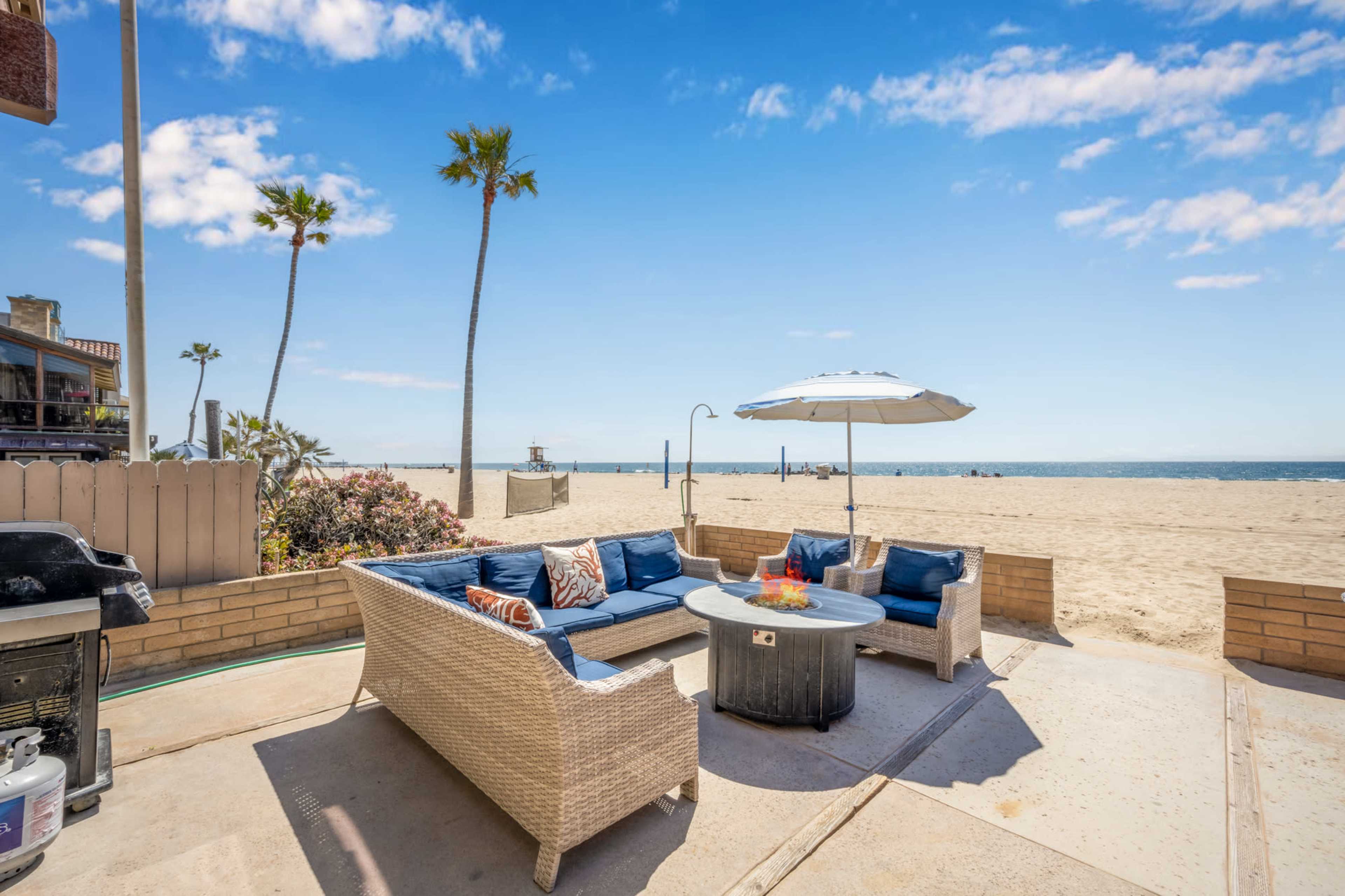 A patio area with a wicker sofa, a fire pit, and an umbrella overlooks a sandy beach and ocean, with palm trees in the background.