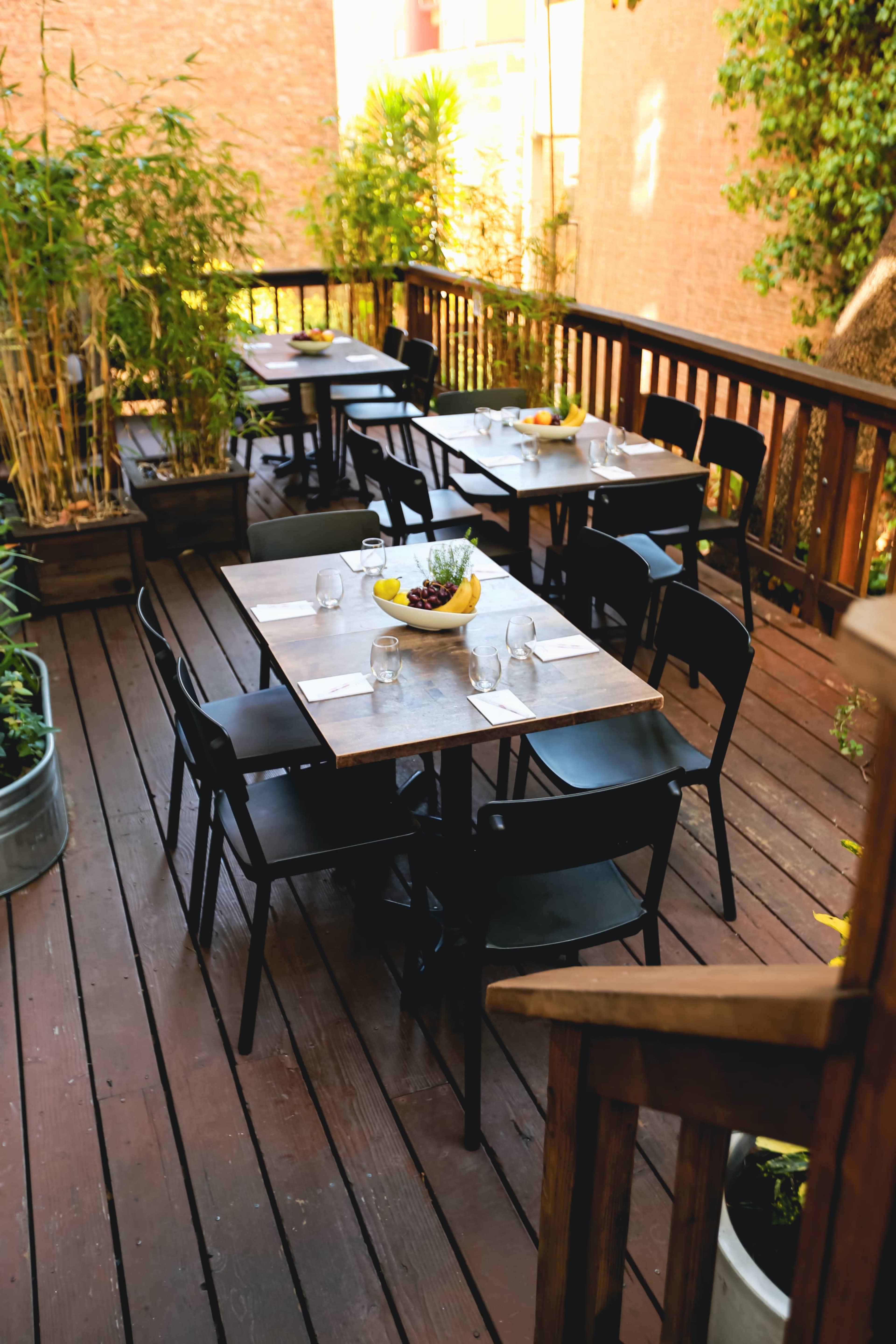 An outdoor dining area with several tables set for meals, surrounded by plants and a wooden railing.