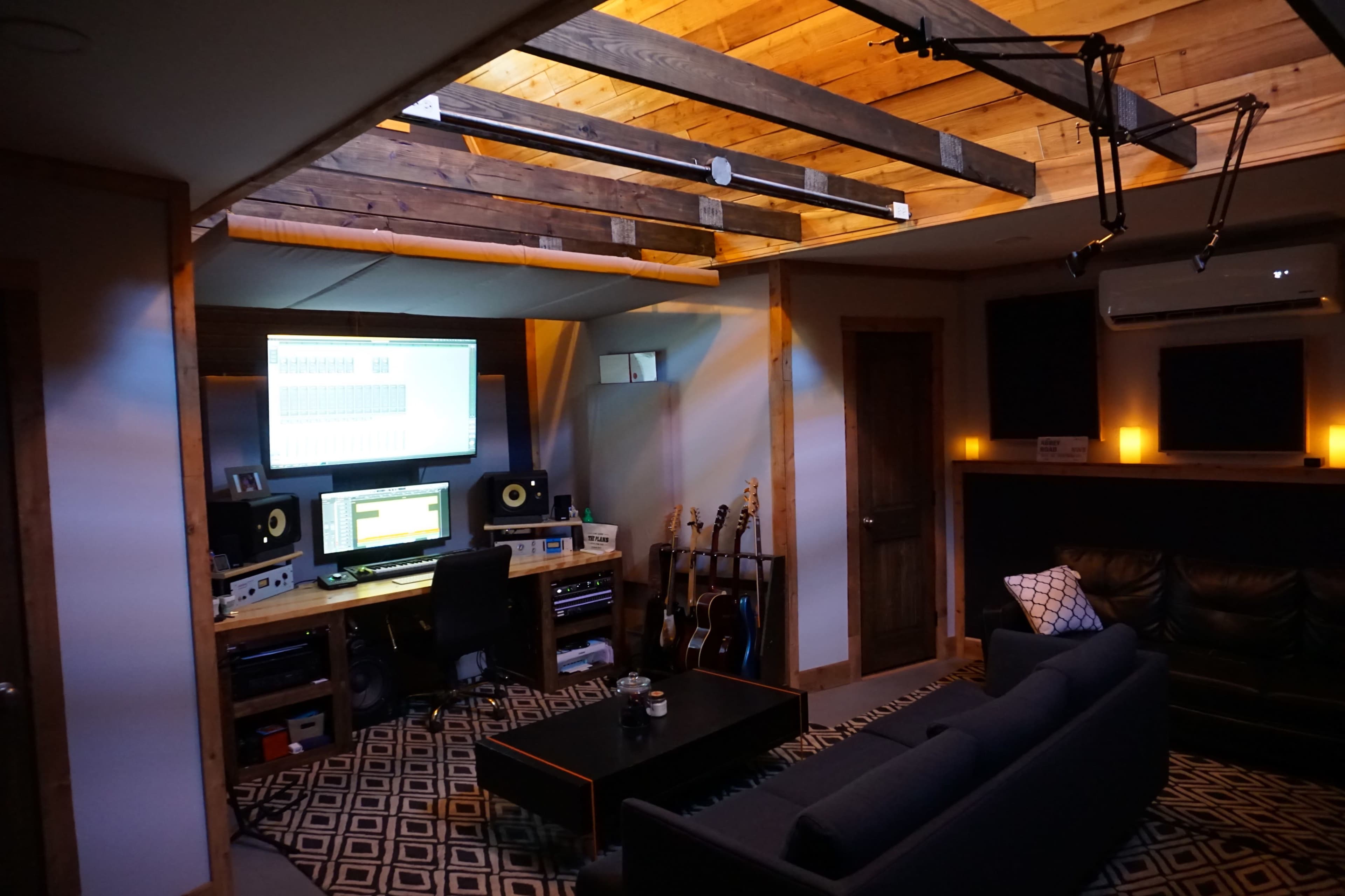 A cozy home recording studio featuring a desk with audio equipment, computer screens, guitars, and a comfortable seating area with a dark sofa.