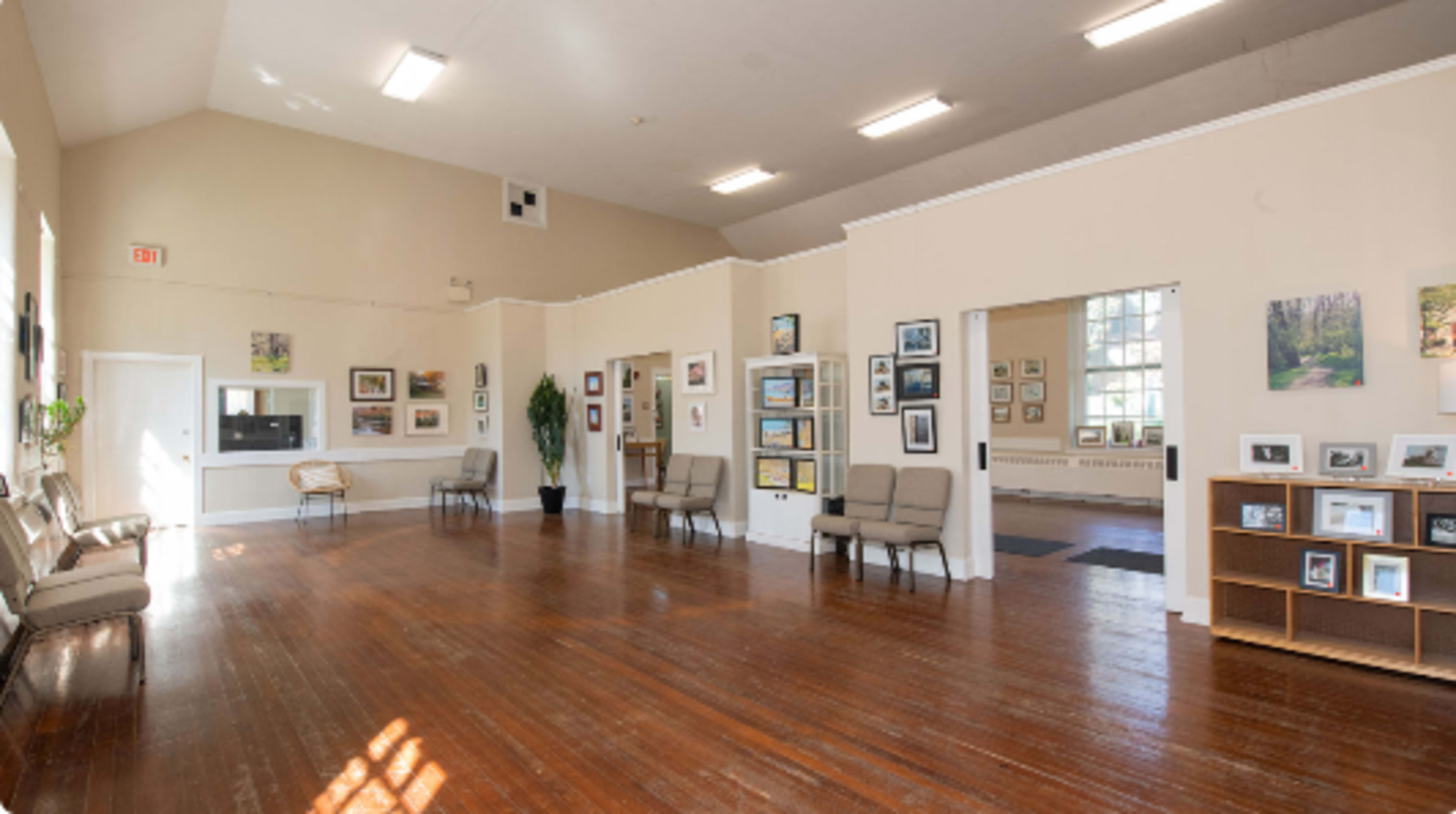 A spacious, well-lit interior room featuring wooden flooring, several framed pictures on the walls, and a mix of seating along one side.