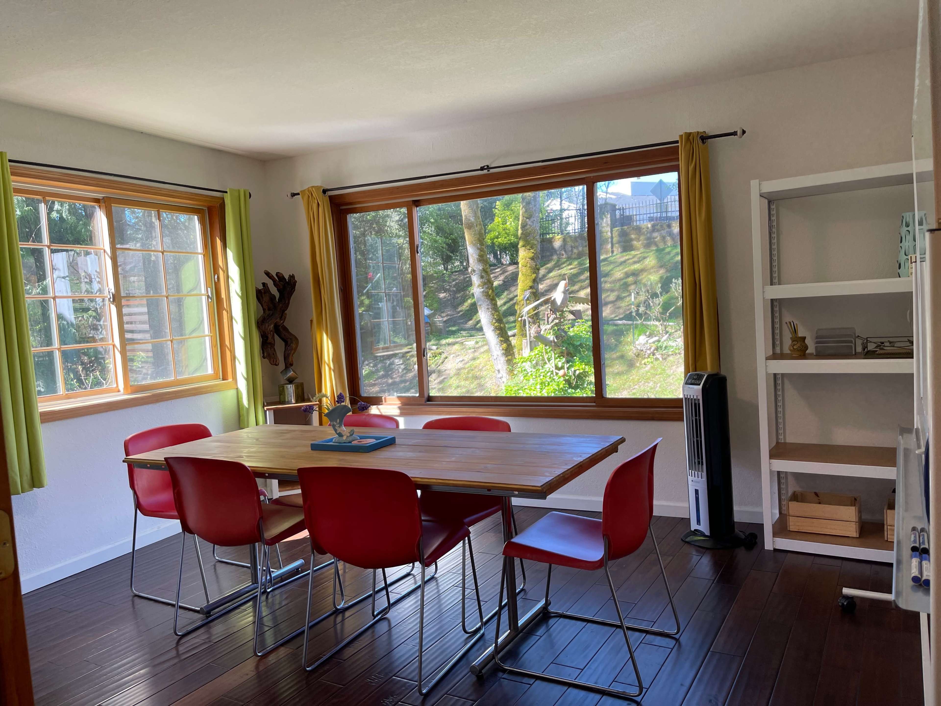 A dining area features a wooden table surrounded by red chairs, with large windows overlooking a green landscape.