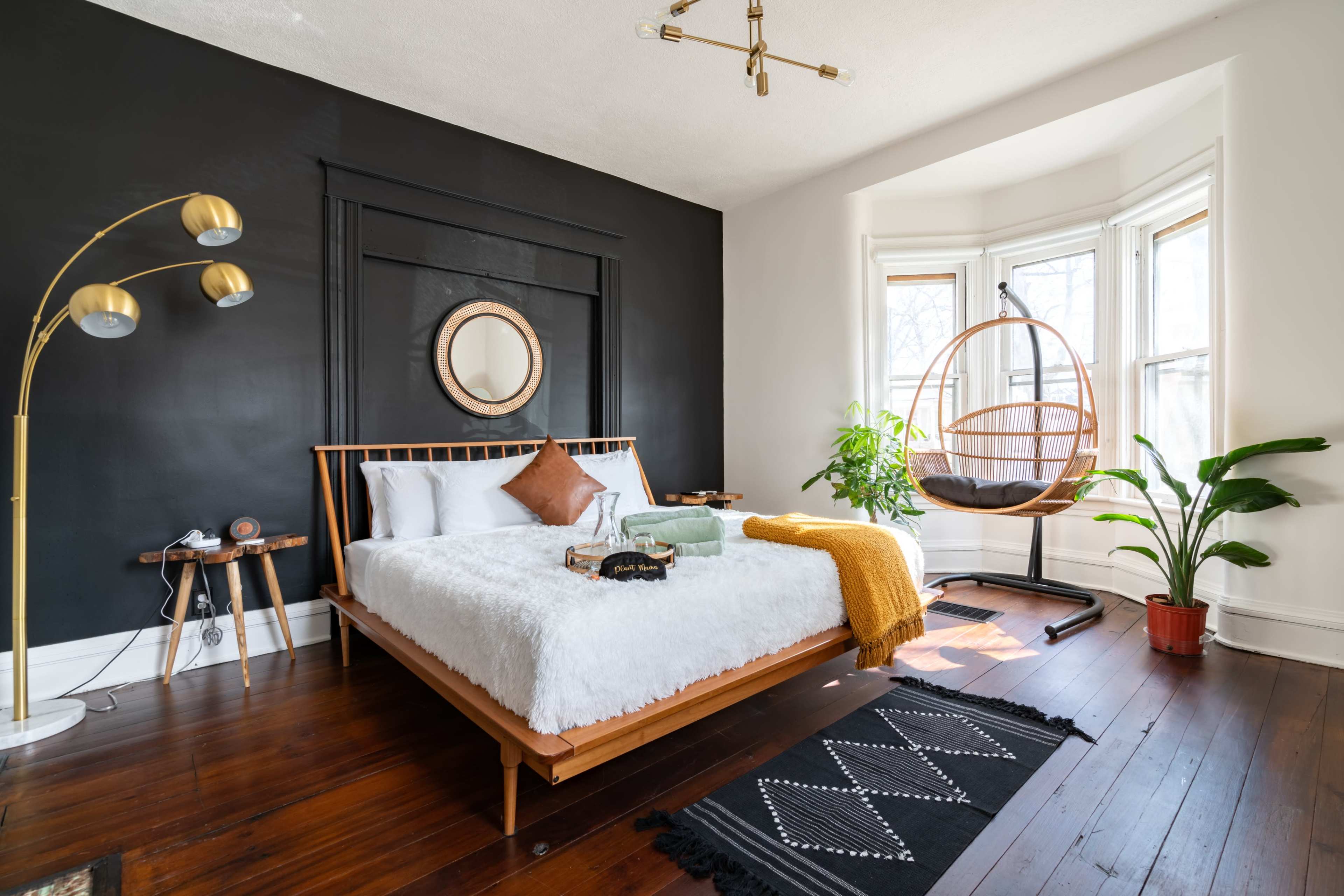 A modern bedroom features a wooden bed with white bedding, a round mirror, a hanging chair by a bay window, and a floor lamp.