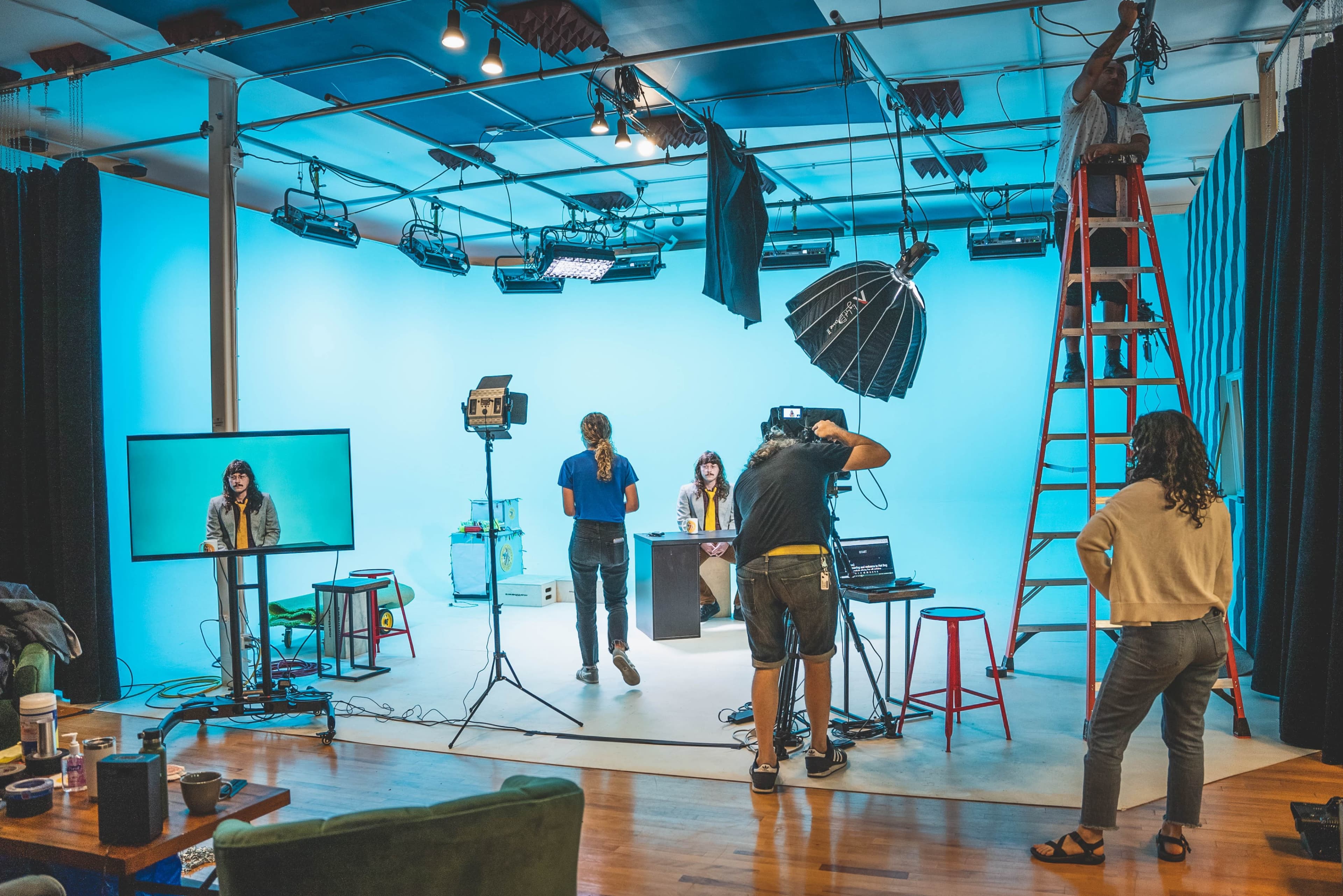 A film crew sets up equipment in a studio with a blue backdrop while a woman sits at a table, and another person films her.