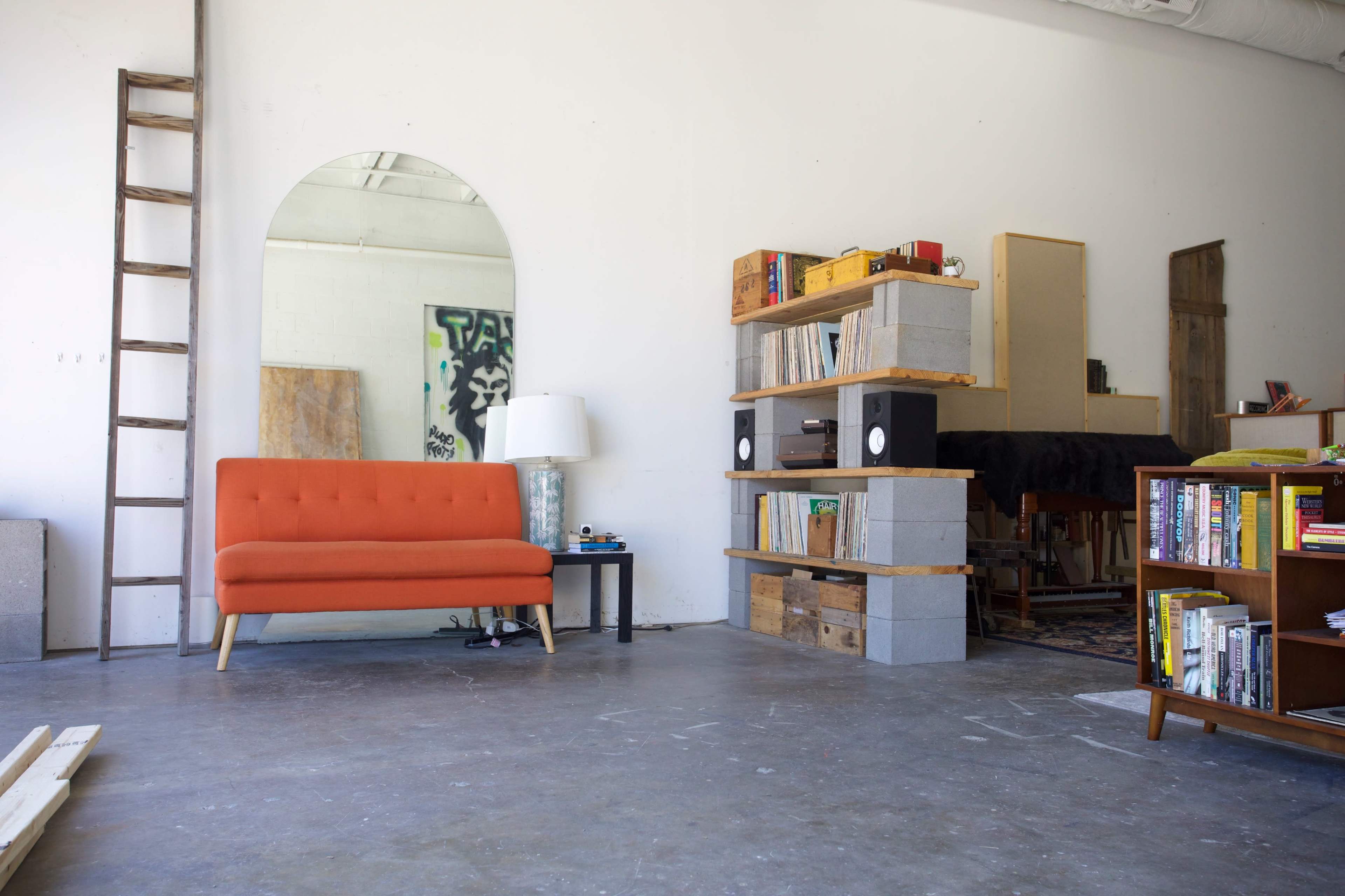 A minimalist interior space featuring an orange sofa, a large mirror, wooden shelves with books and speakers, and a ladder leaning against the wall.