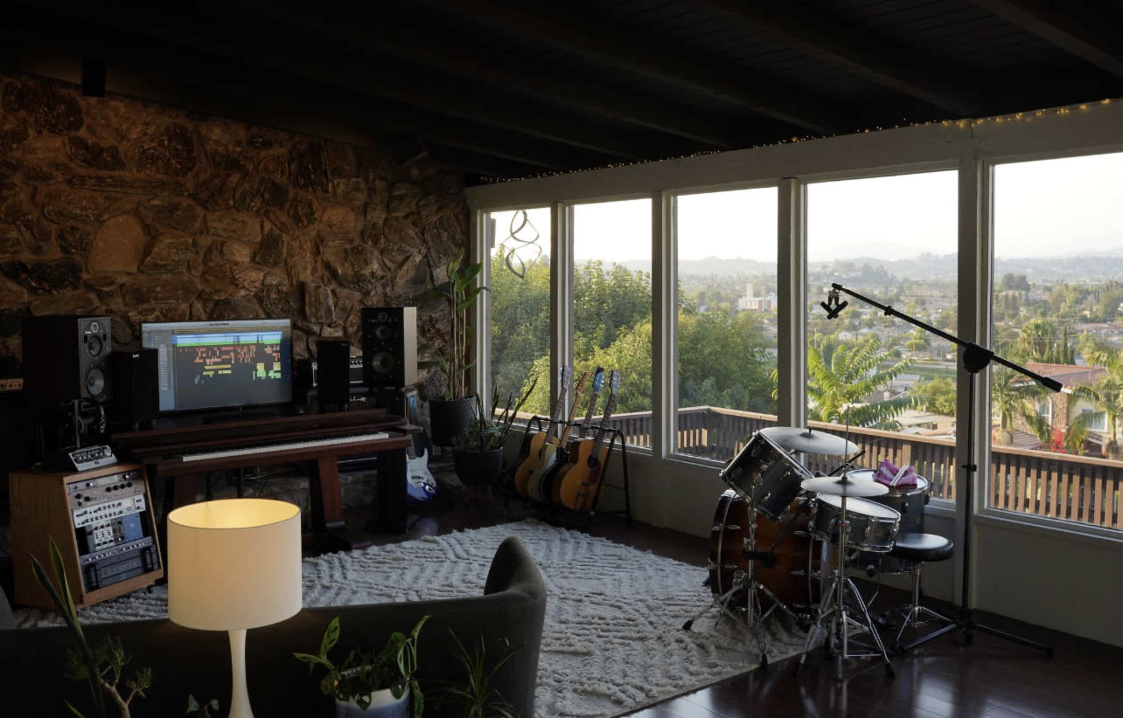 A spacious room features a stone wall, a piano, speakers, a drum set, and large windows with a view of a landscape.