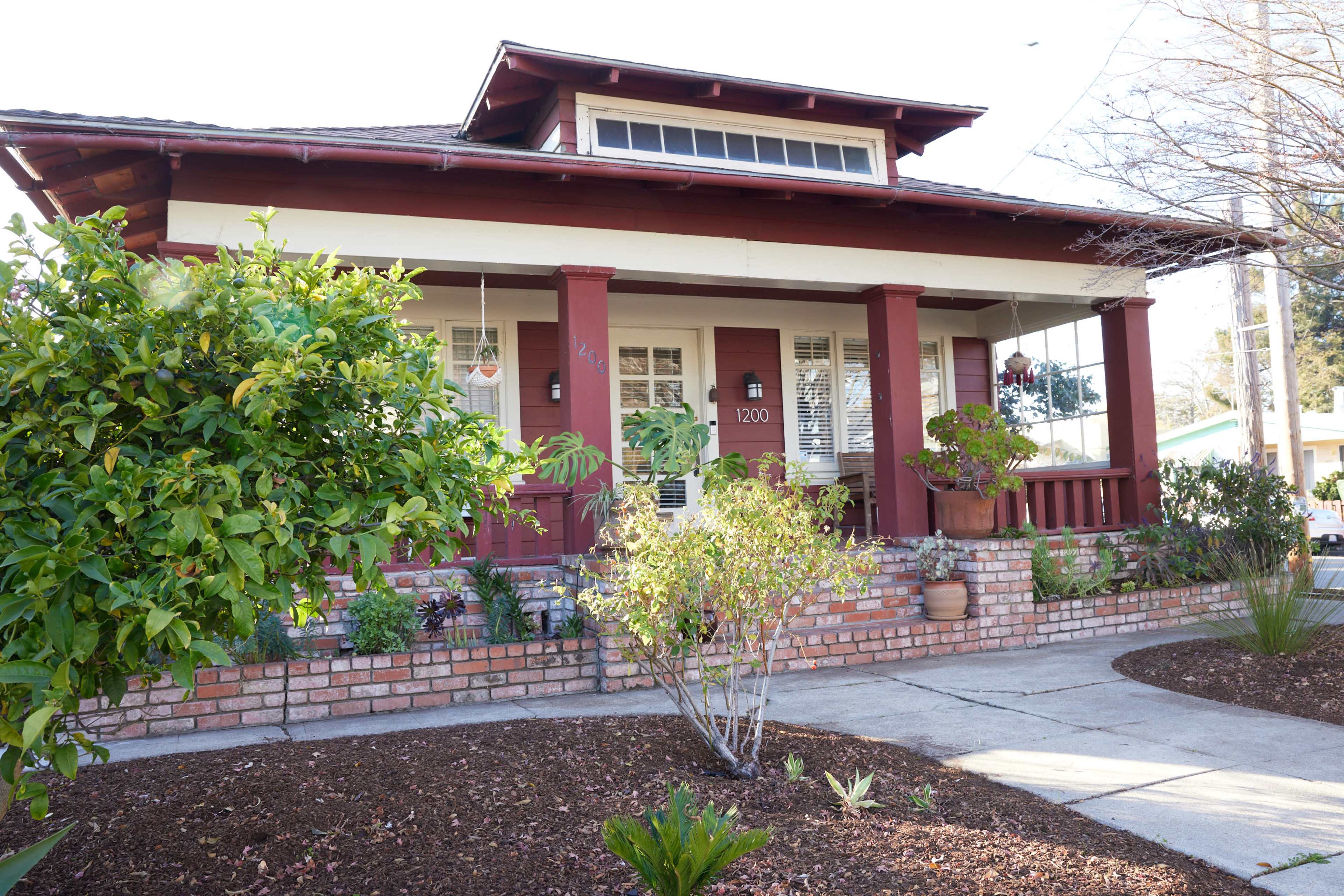A bungalow-style house with a red facade, a front porch supported by thick columns, and a landscaped yard with various plants and shrubs.