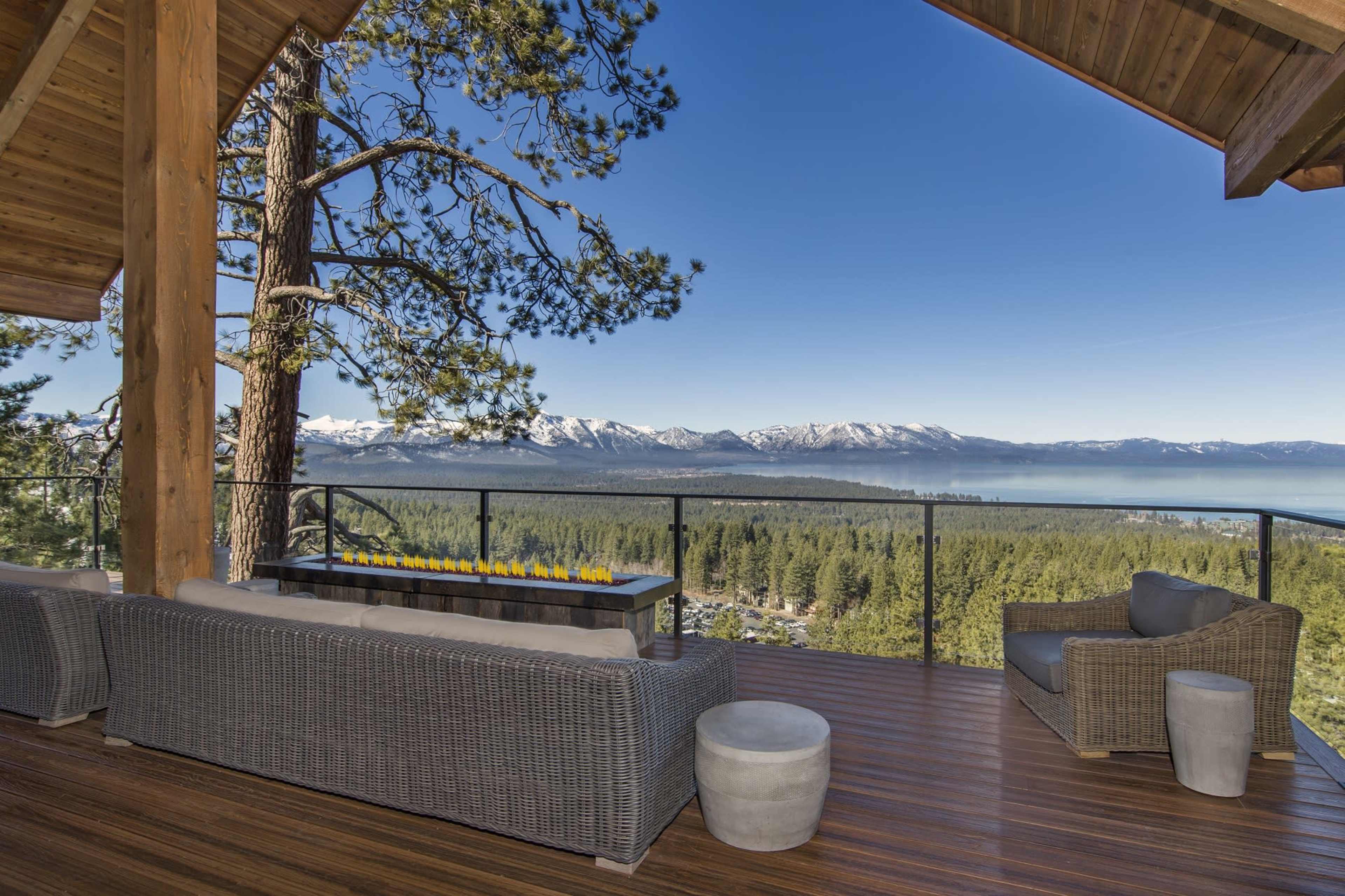 A spacious deck features comfortable seating and a fire table, overlooking a scenic view of mountains and a lake surrounded by trees.