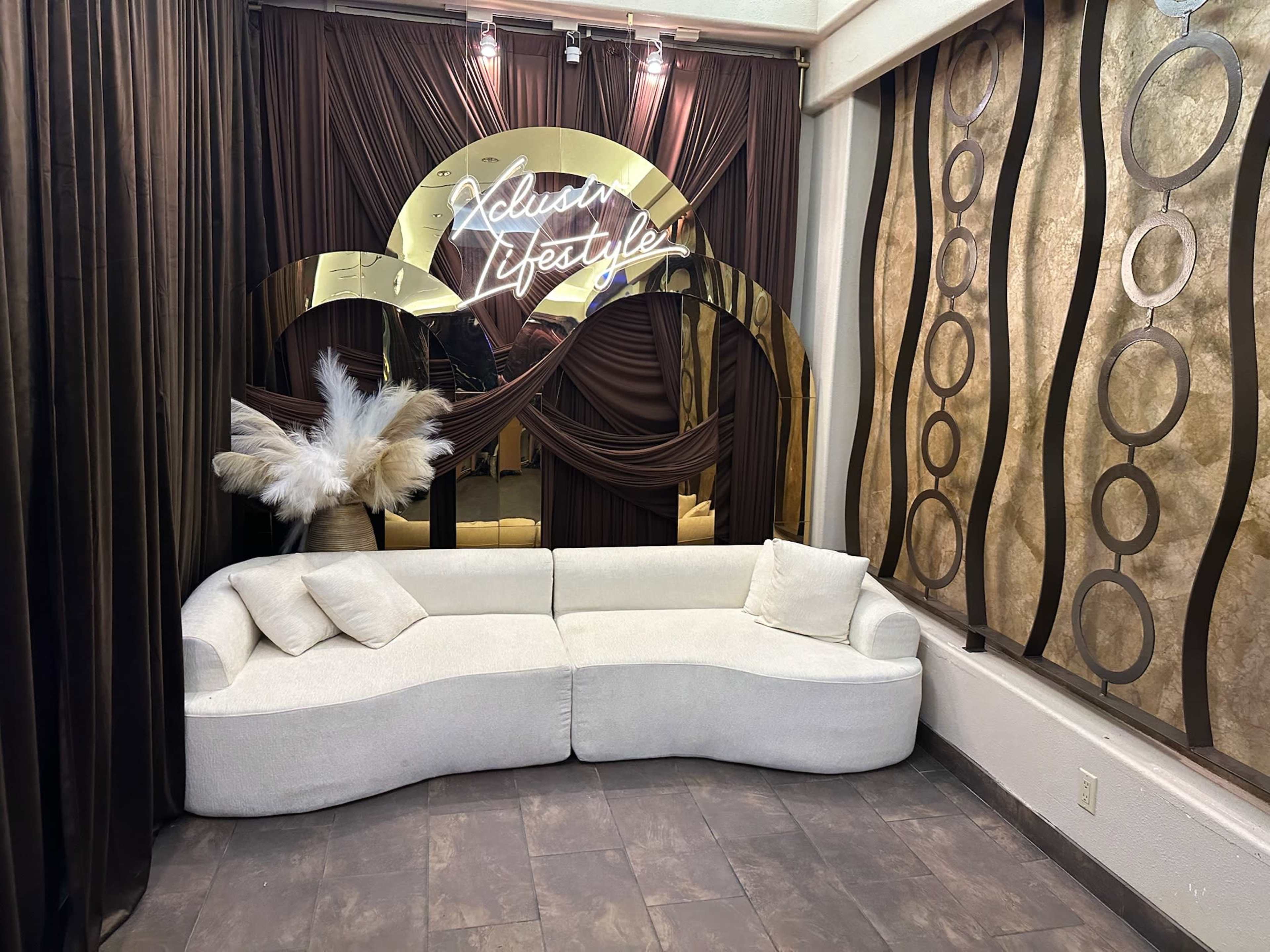 The image shows a modern, curved white couch positioned beneath a decorative wall with brown drapery and a neon sign that reads "Xclusive Lifestyle."