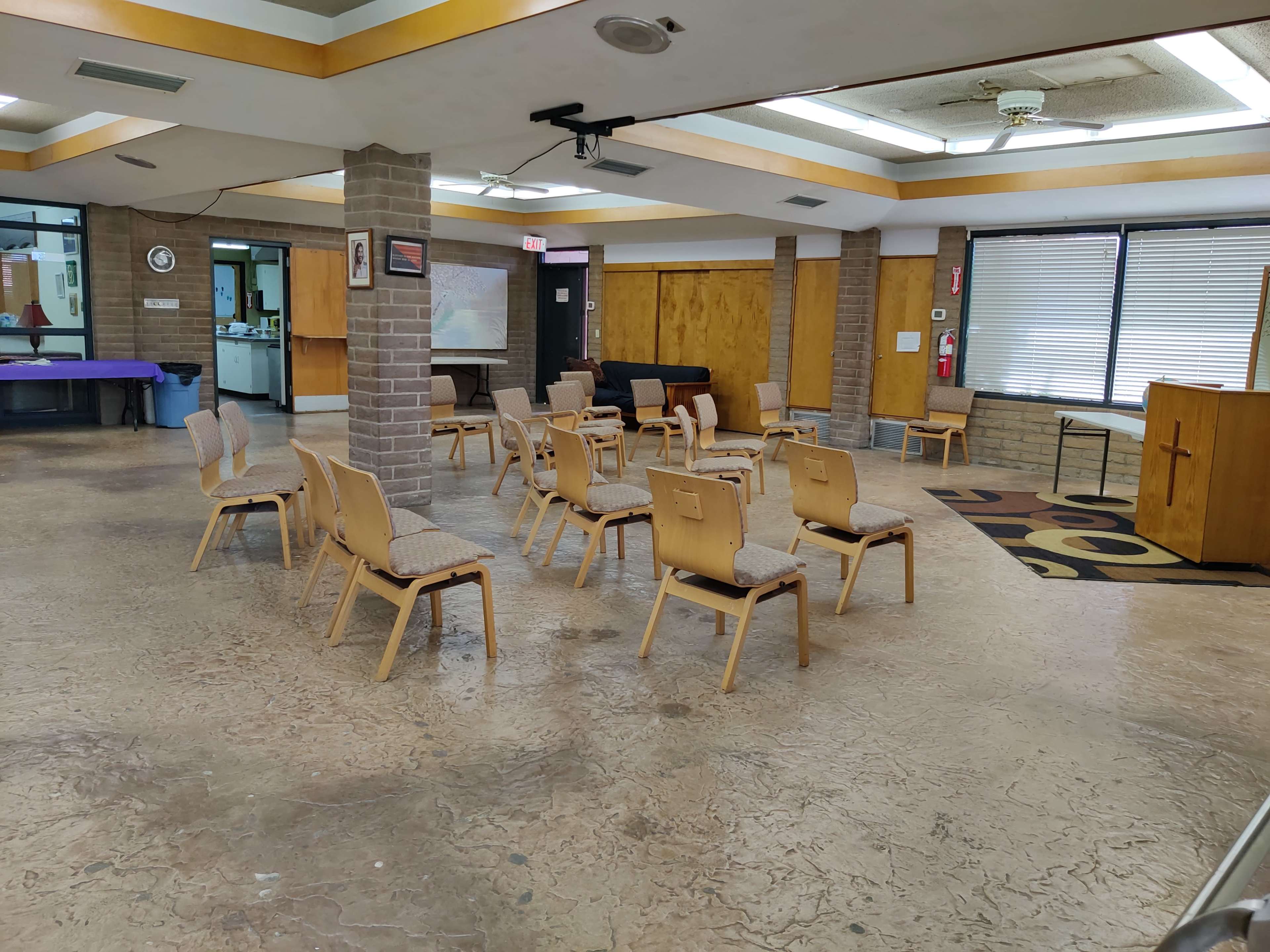 A room with several wooden chairs arranged in rows around a central space, featuring a podium with a cross and large windows along one side.