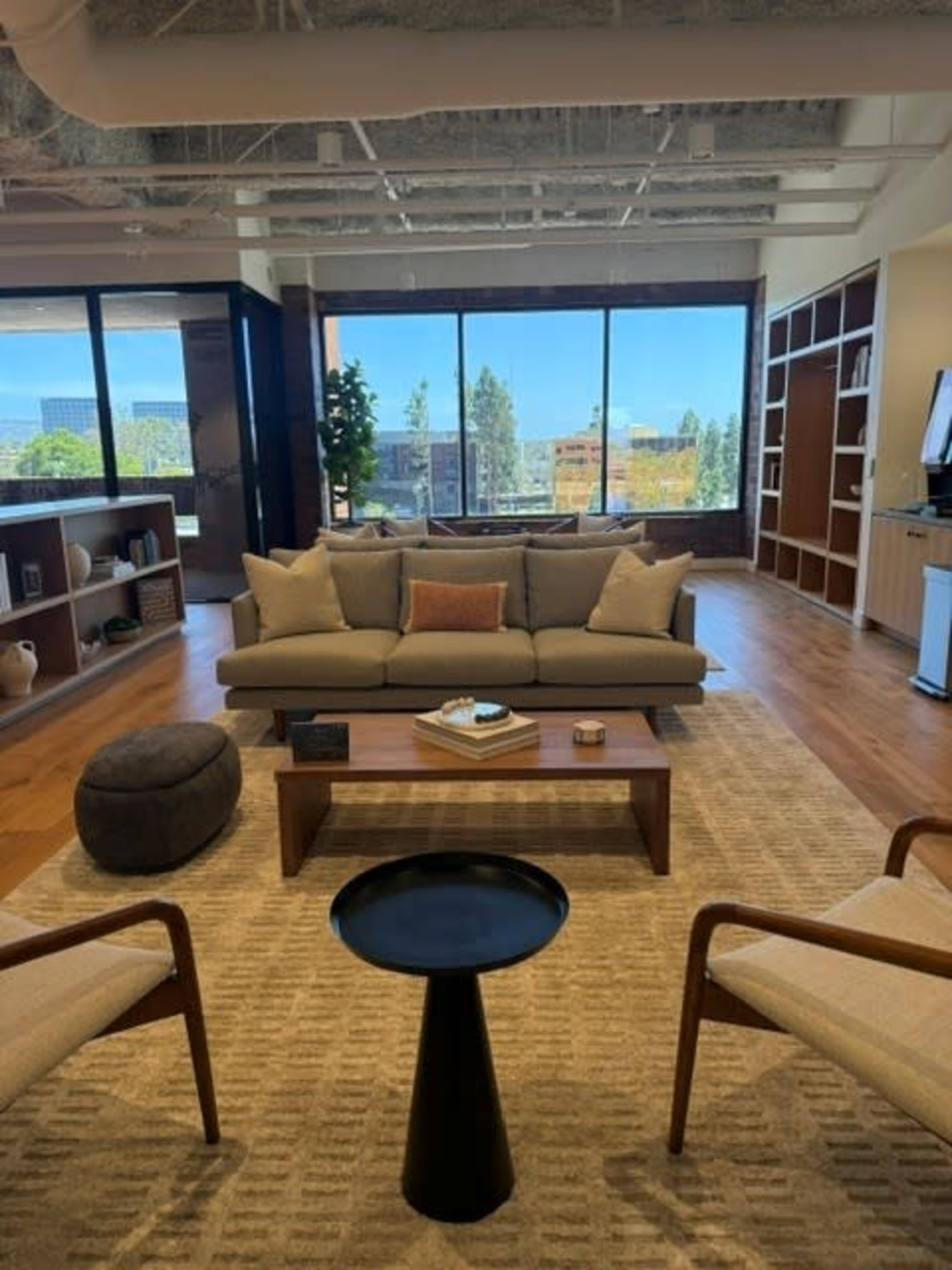 The image shows a modern office lounge area featuring a sofa, coffee table, and armchairs, with large windows displaying a view of the outdoors.