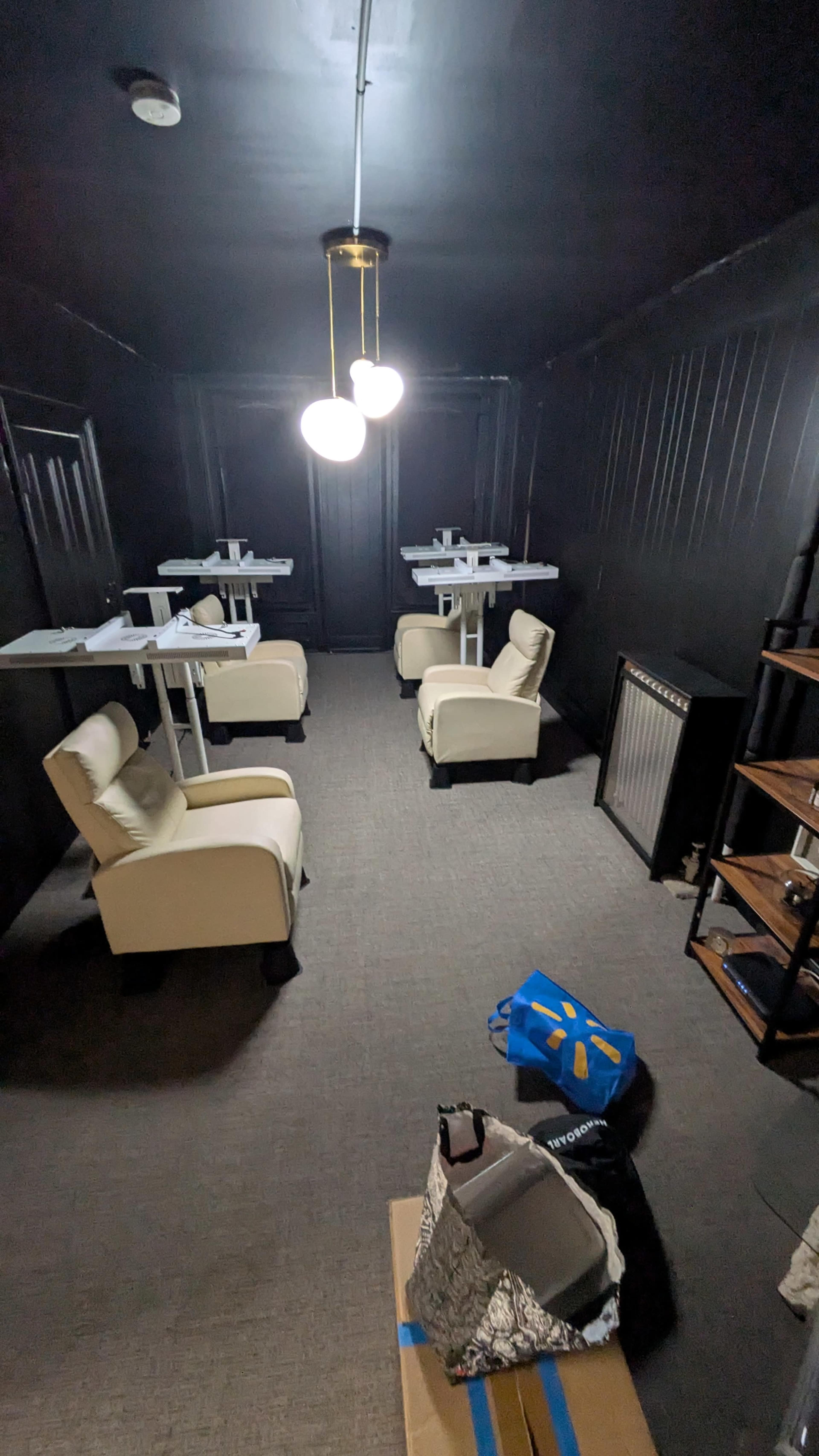 The image shows a dimly lit room with black walls, featuring four white tables with chairs, a shelf, and some bags on the floor.