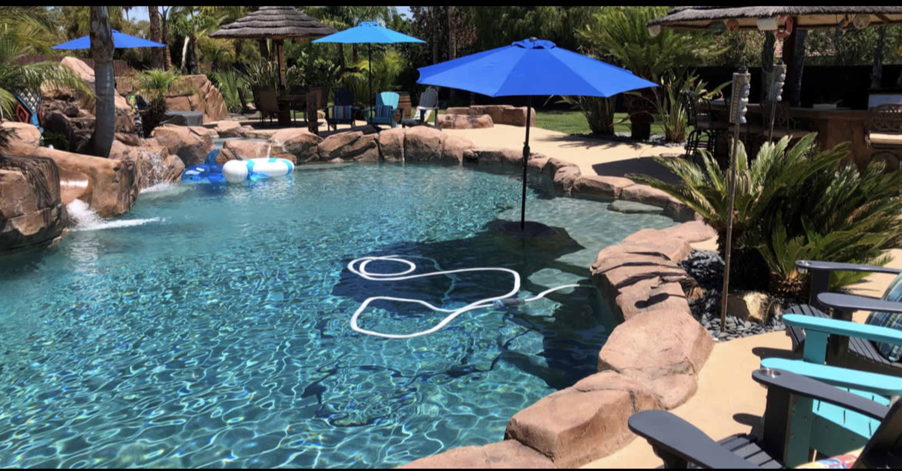 A landscaped swimming pool area with rocky formations, lounge chairs, umbrellas, and a waterfall feature.