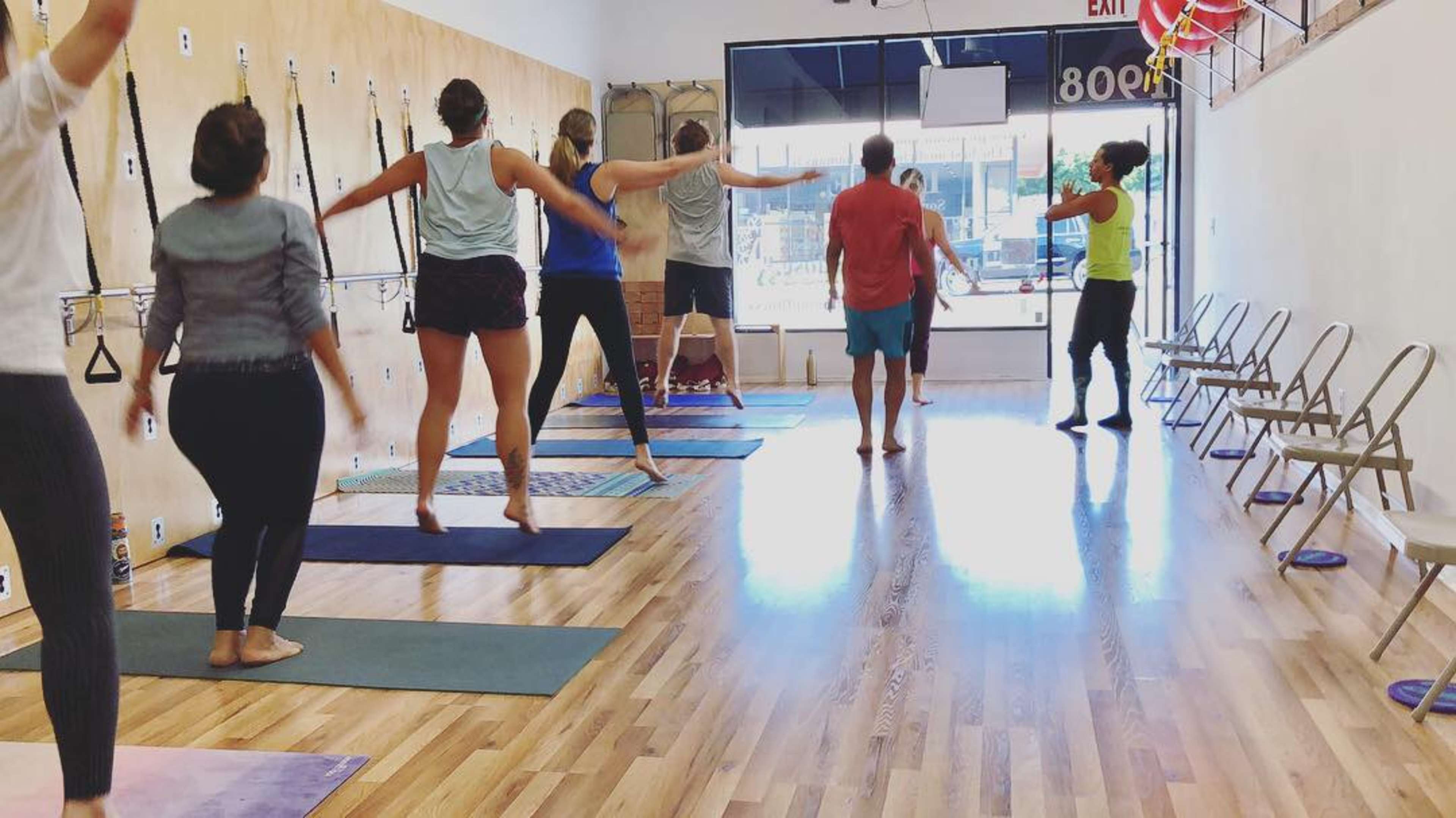 A group of people is participating in a fitness class inside a studio with yoga mats and equipment against the wall.