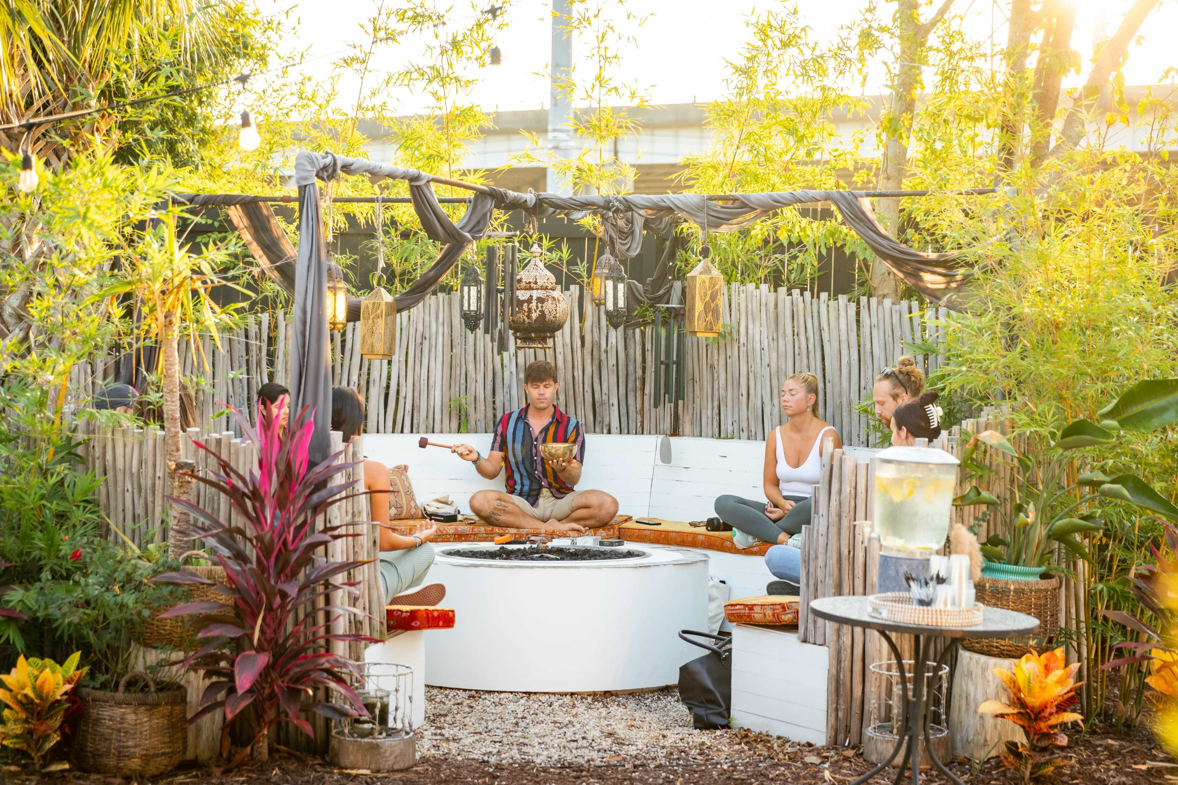 A group of individuals sits in a circular arrangement around a central fire pit in a cozy outdoor space surrounded by greenery and wooden fencing.