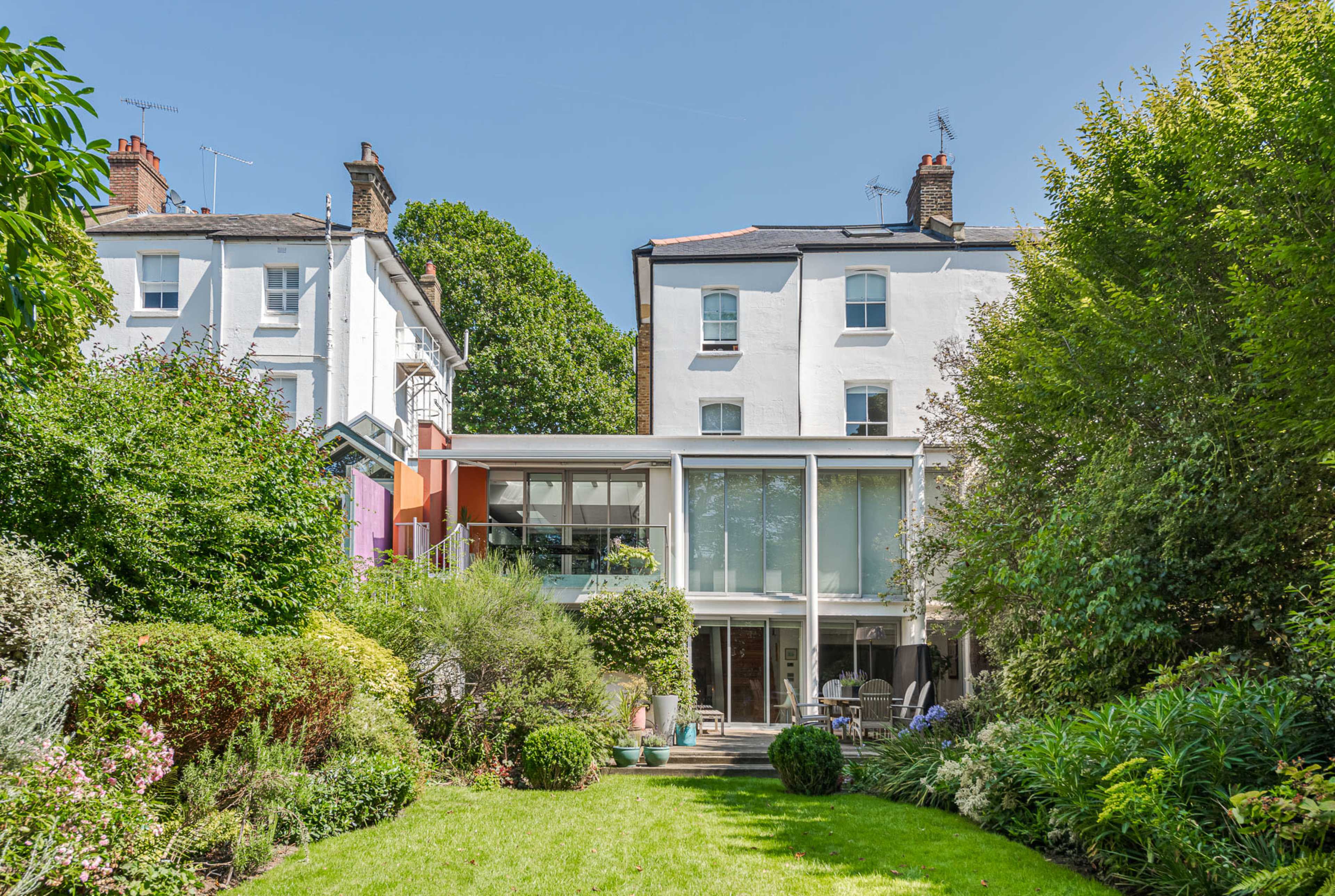 A modern glass-walled house sits at the end of a lush garden surrounded by greenery and neighboring buildings.