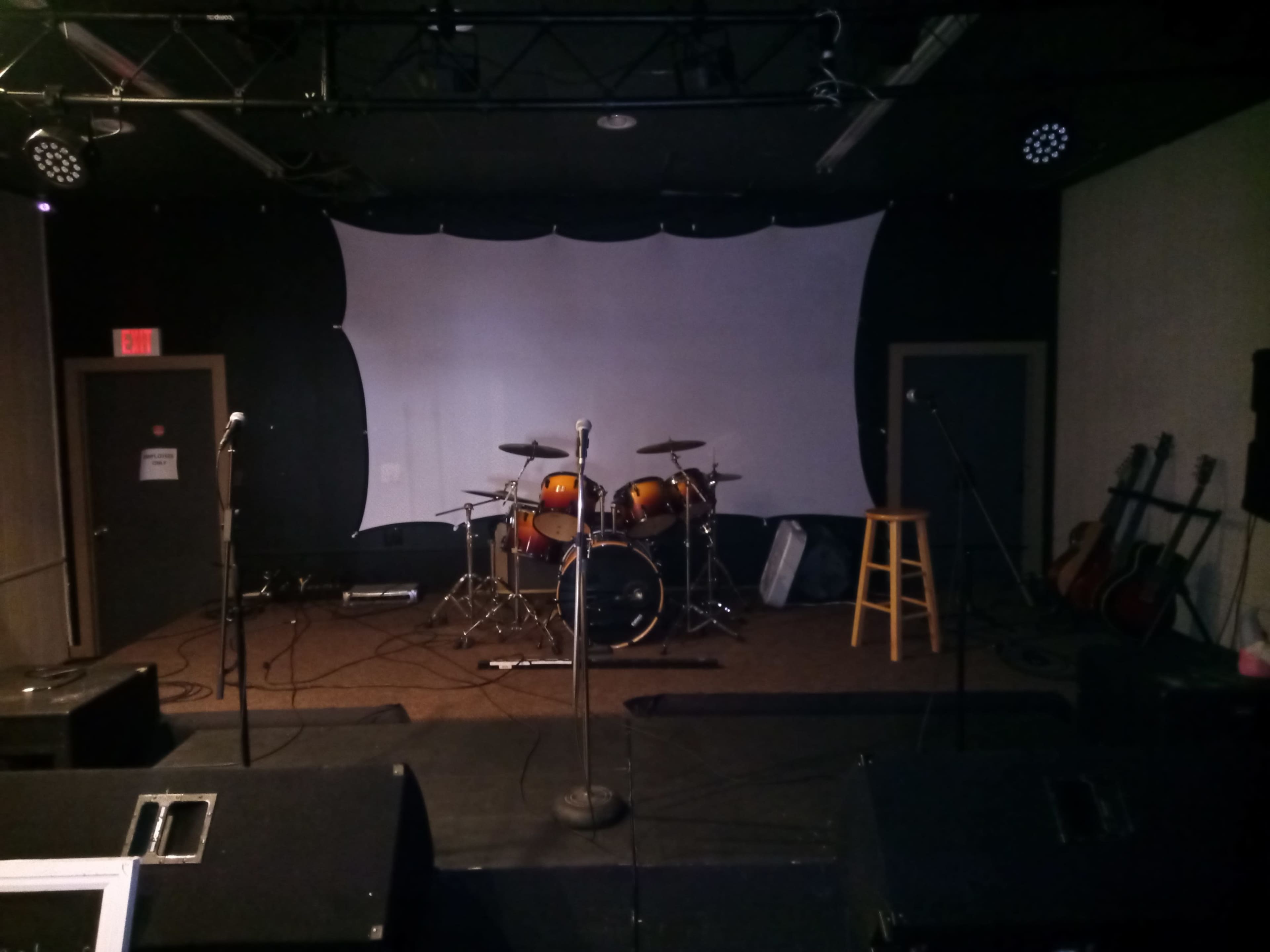 A performance stage featuring a drum set, microphones, a stool, and guitars against a backdrop with a white screen.