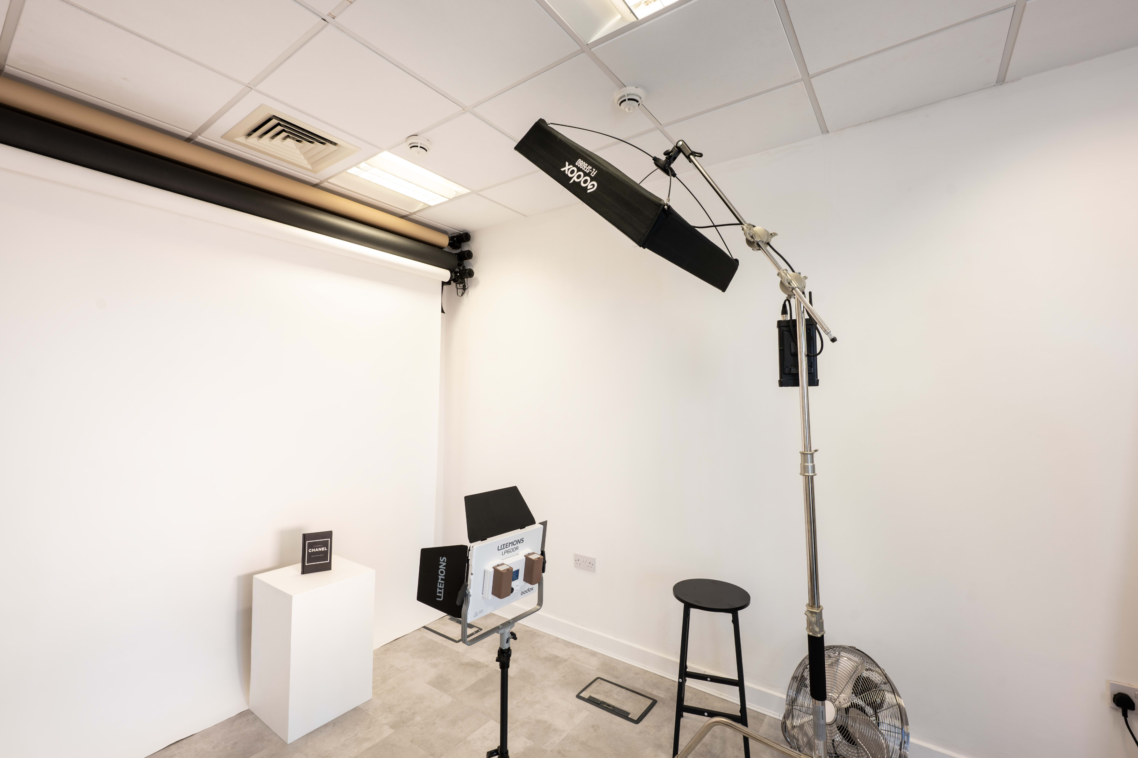 The image shows a minimalistic photography studio featuring a light setup, a stool, a pedestal for displays, and a fan against a white wall.