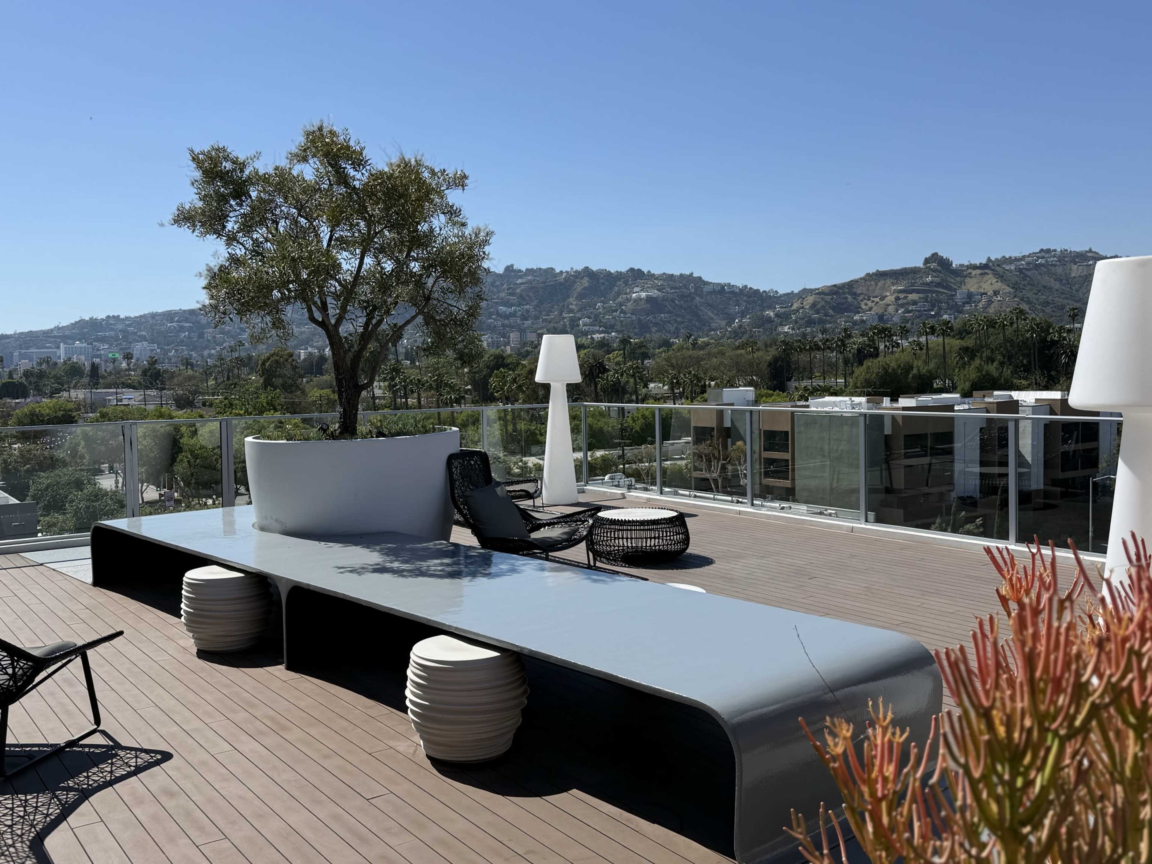 A spacious rooftop terrace features a modern seating area, a large table, and potted plants, with a view of distant hills and a clear blue sky.