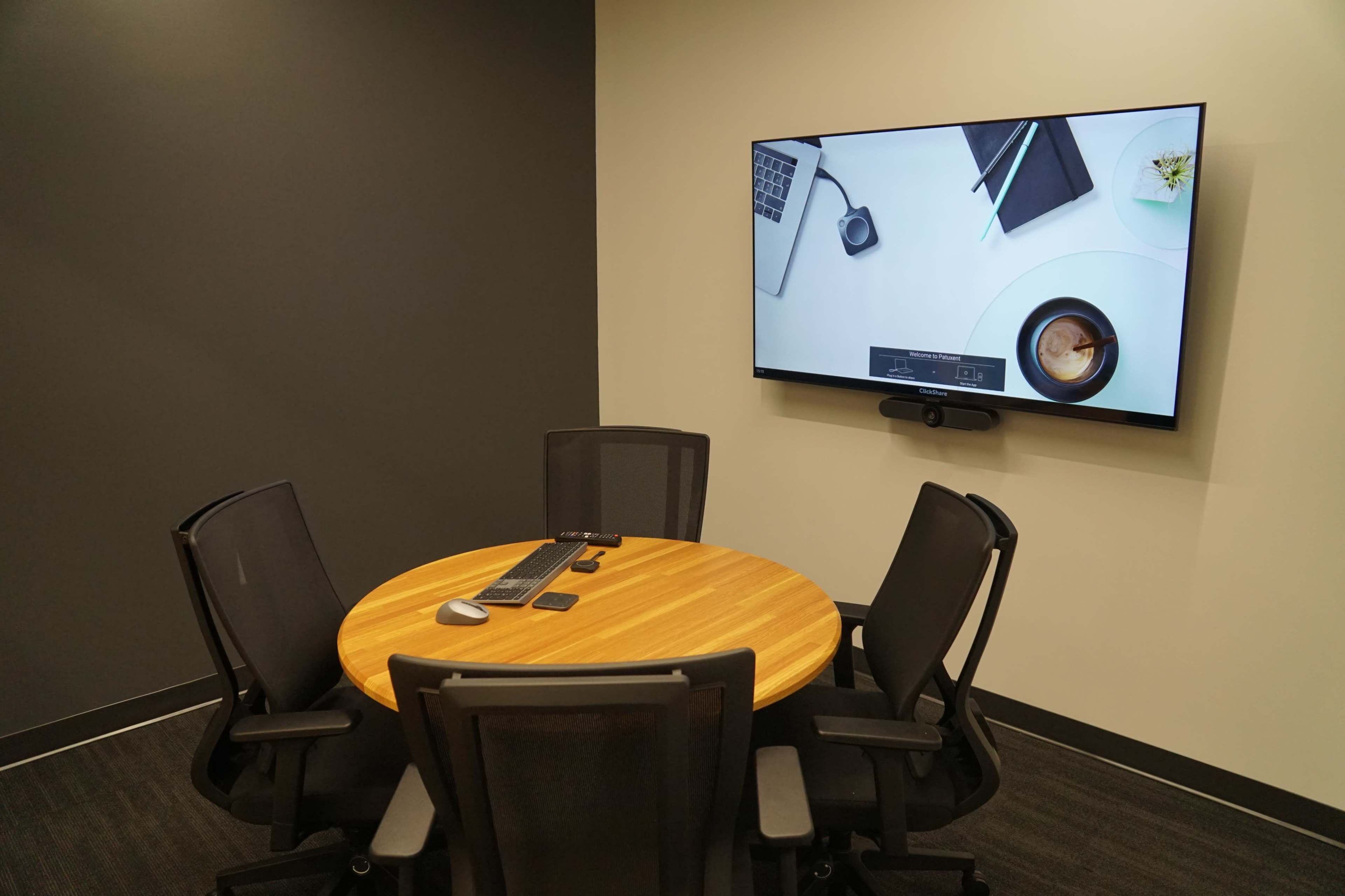 A small meeting room features a round wooden table with four black chairs and a wall-mounted screen displaying a laptop and a coffee cup.