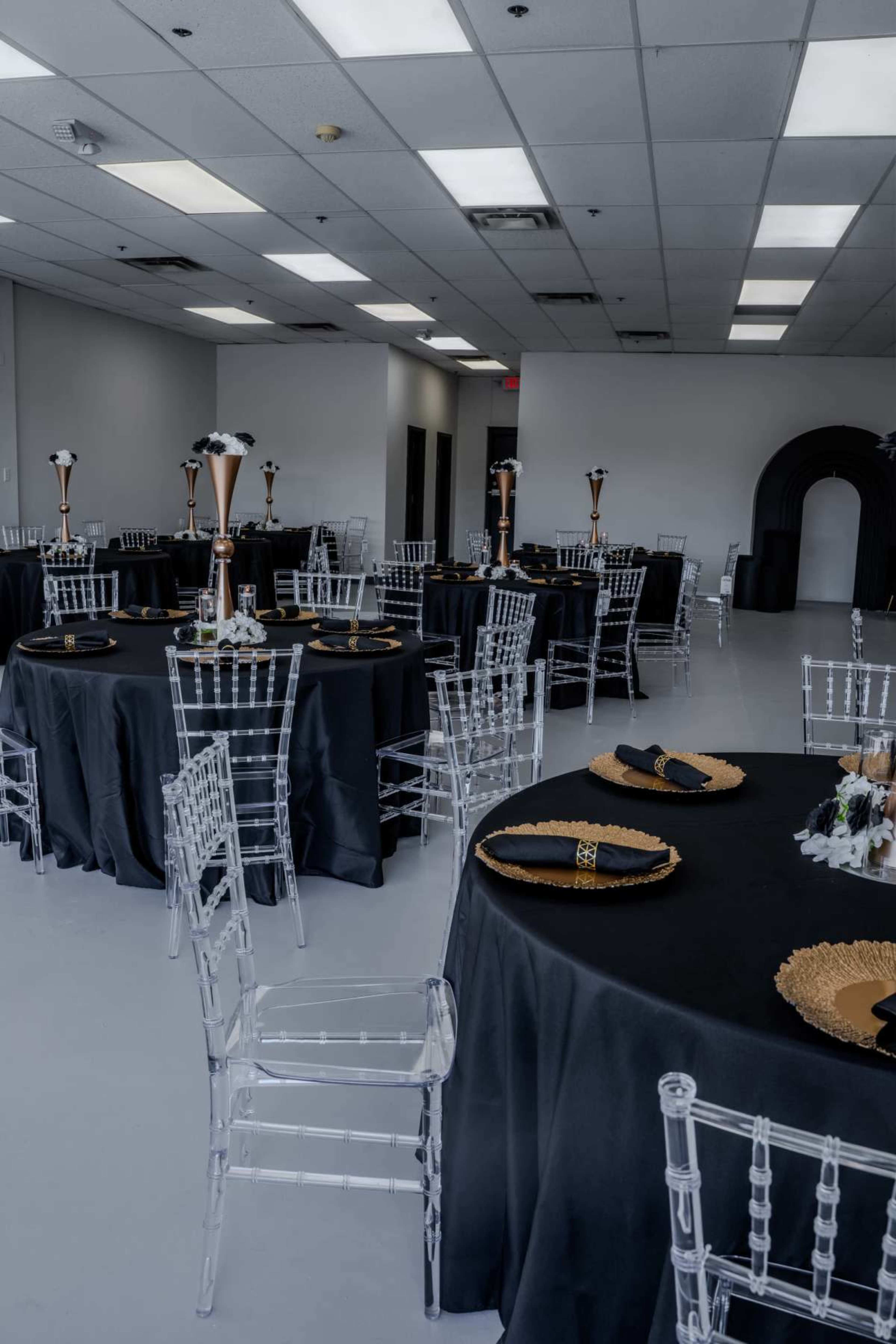 A banquet hall is set with black tablecloths, gold-accented plates, and clear ghost chairs arranged around several round tables.