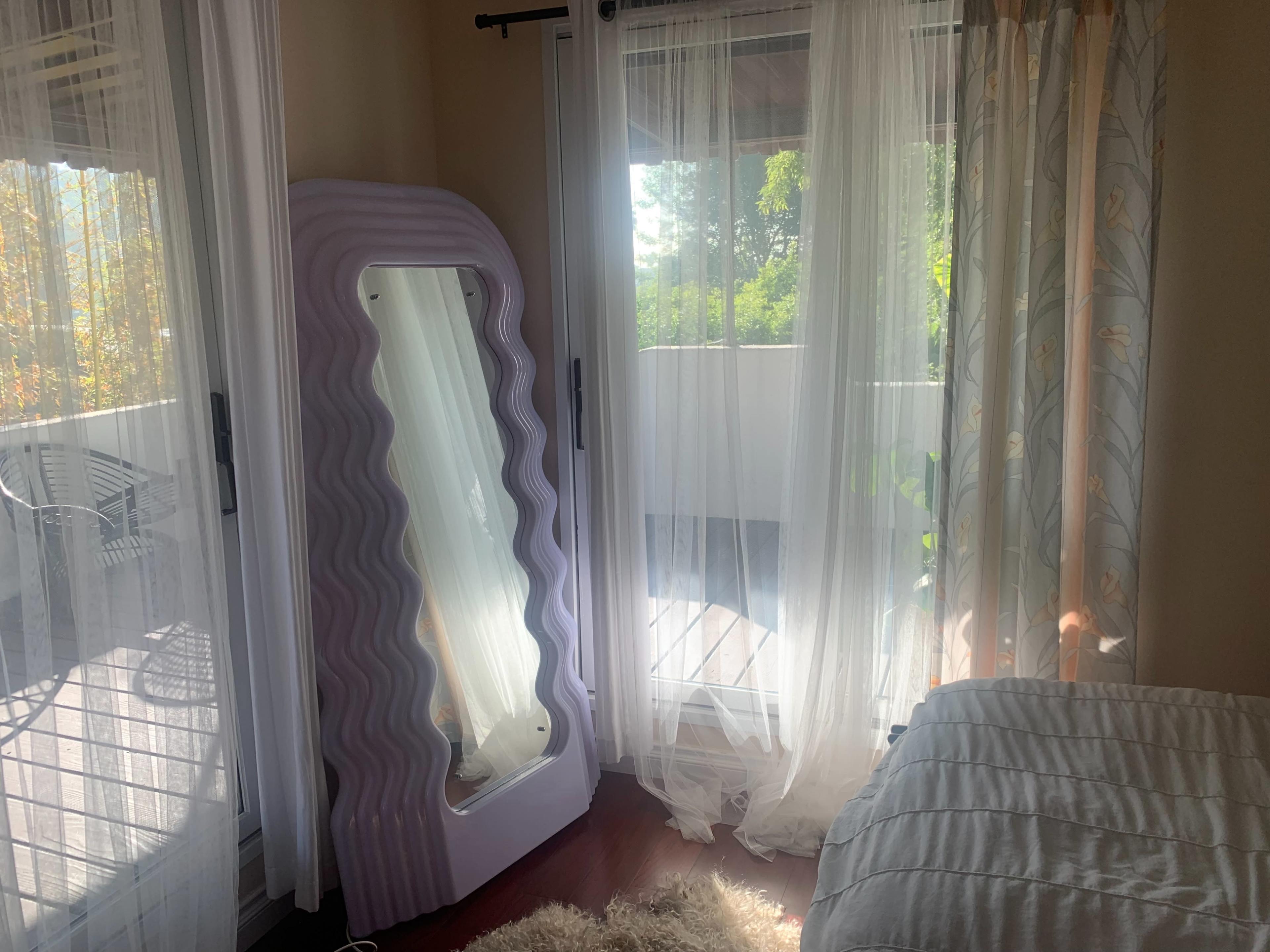 A tall, wavy mirror stands beside sheer curtains that frame a bright, sunlit window in a bedroom.