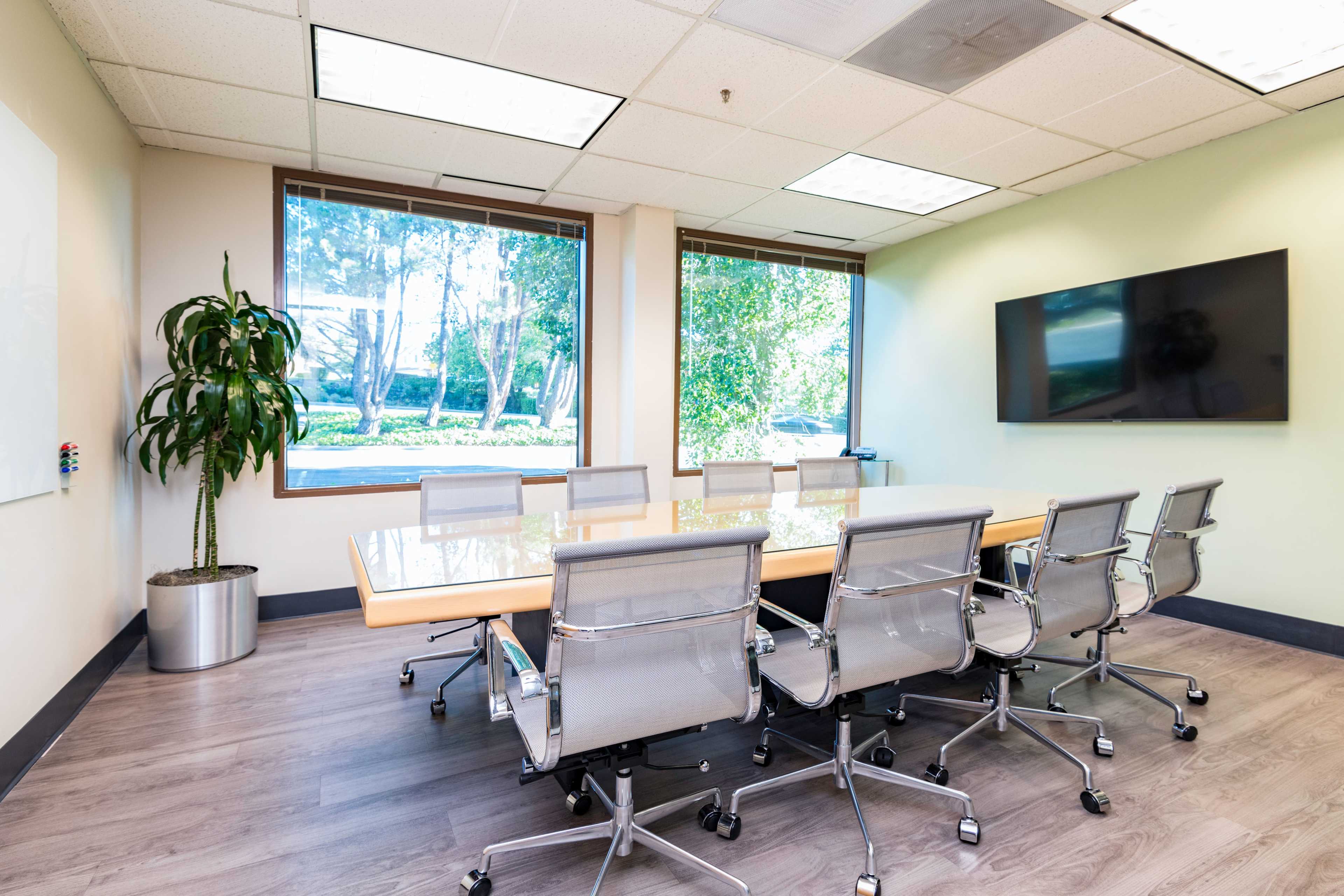 A conference room features a long table surrounded by several chairs, with large windows offering a view of greenery outside.