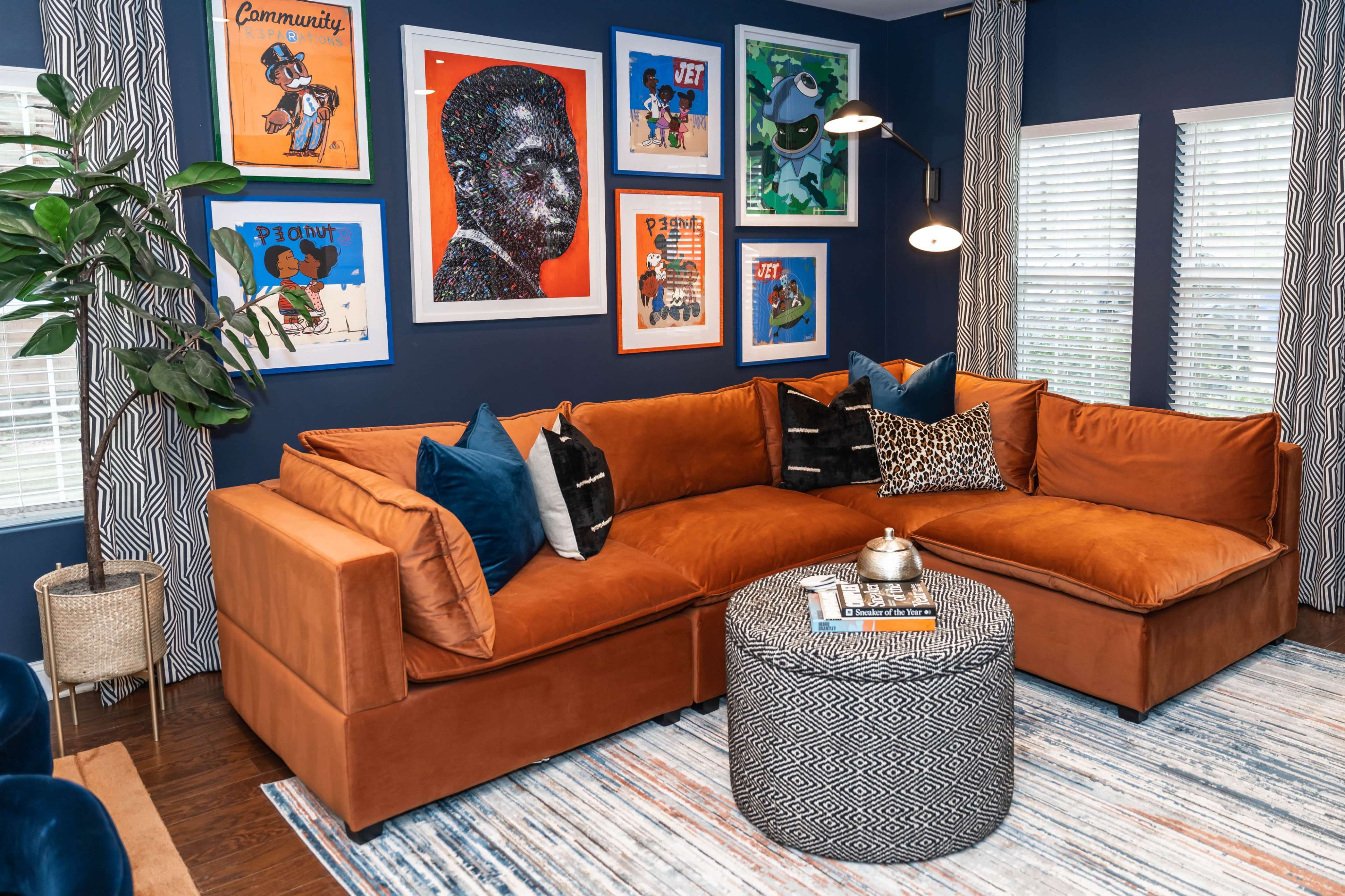 The room features a large, brown sectional couch with colorful throw pillows, a patterned round coffee table, and framed artwork on the blue walls.