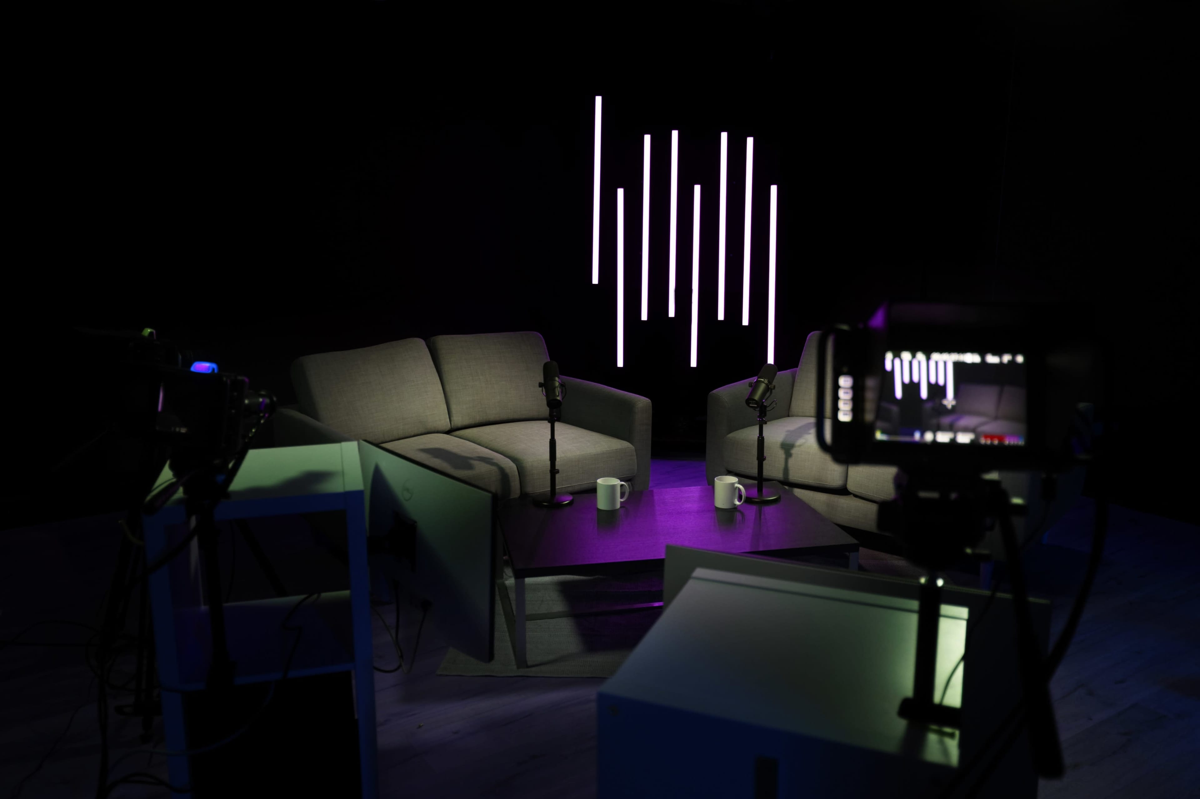 A dimly lit studio features two gray couches facing each other, with microphones and coffee mugs on a small table, alongside a camera and illuminated vertical light strips in the background.