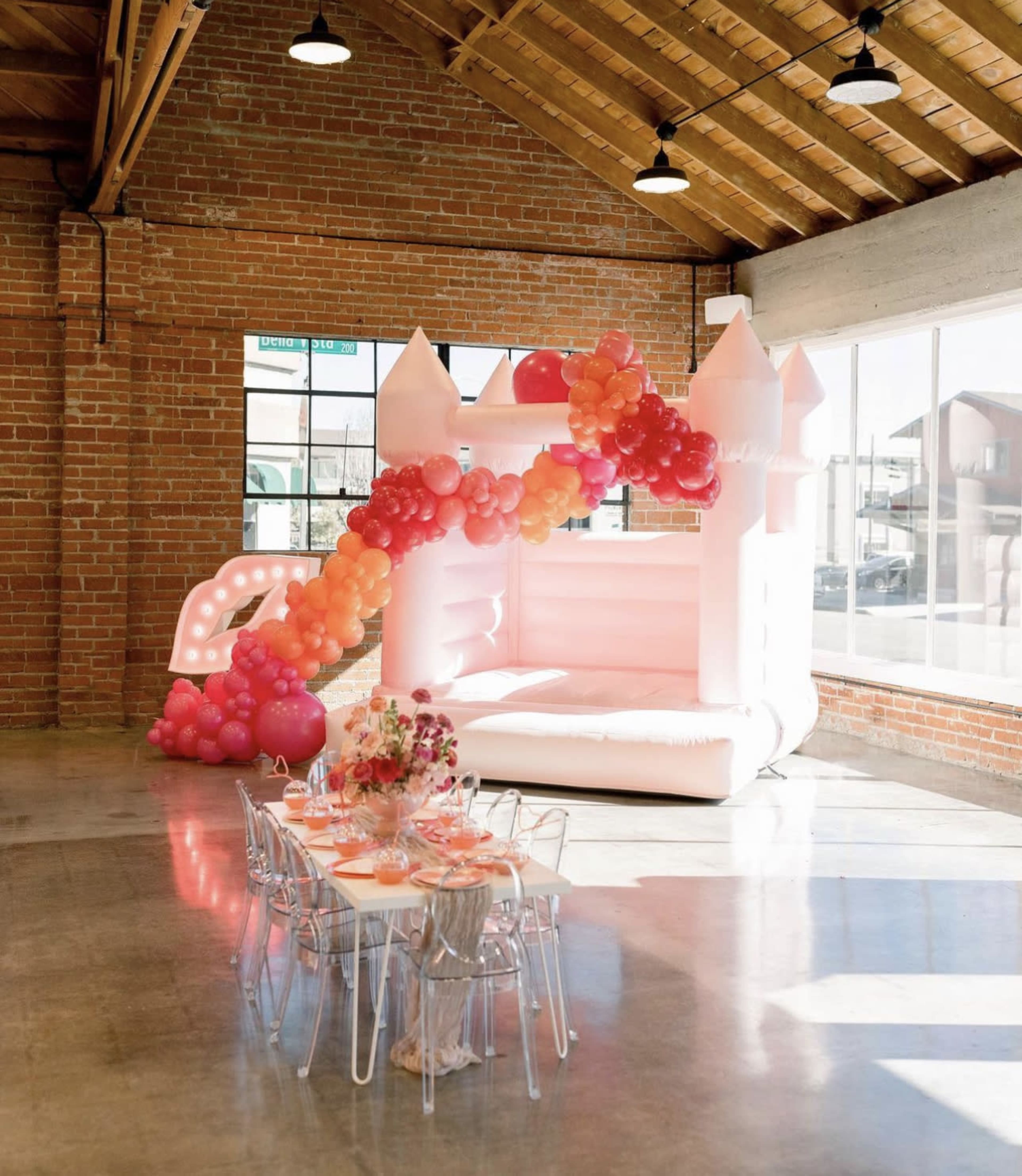 The image shows an indoor space featuring a pink inflatable castle with a balloon arch and a dining table set for a celebration.