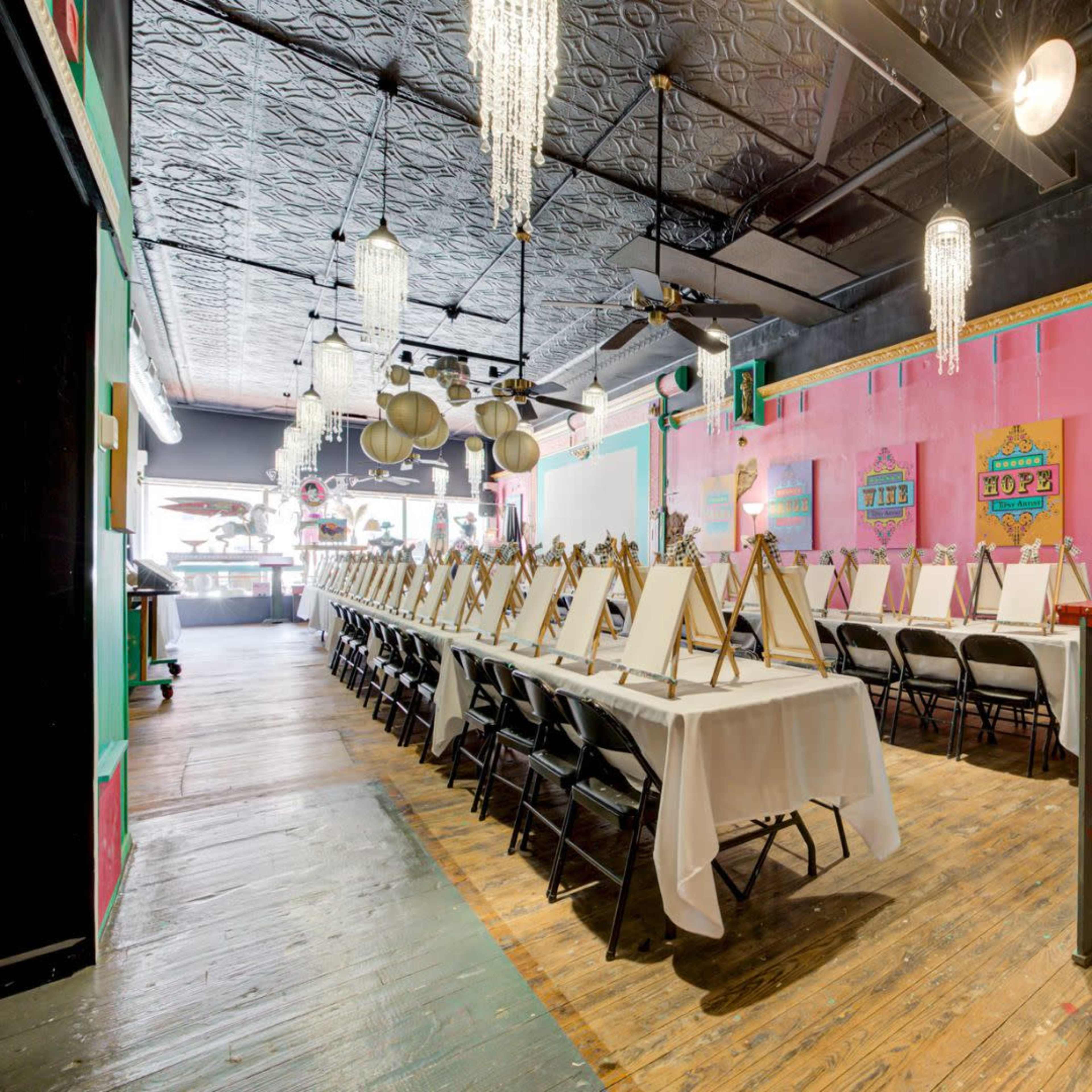 The image shows a brightly decorated art studio with rows of easels and chairs set up for a painting event.