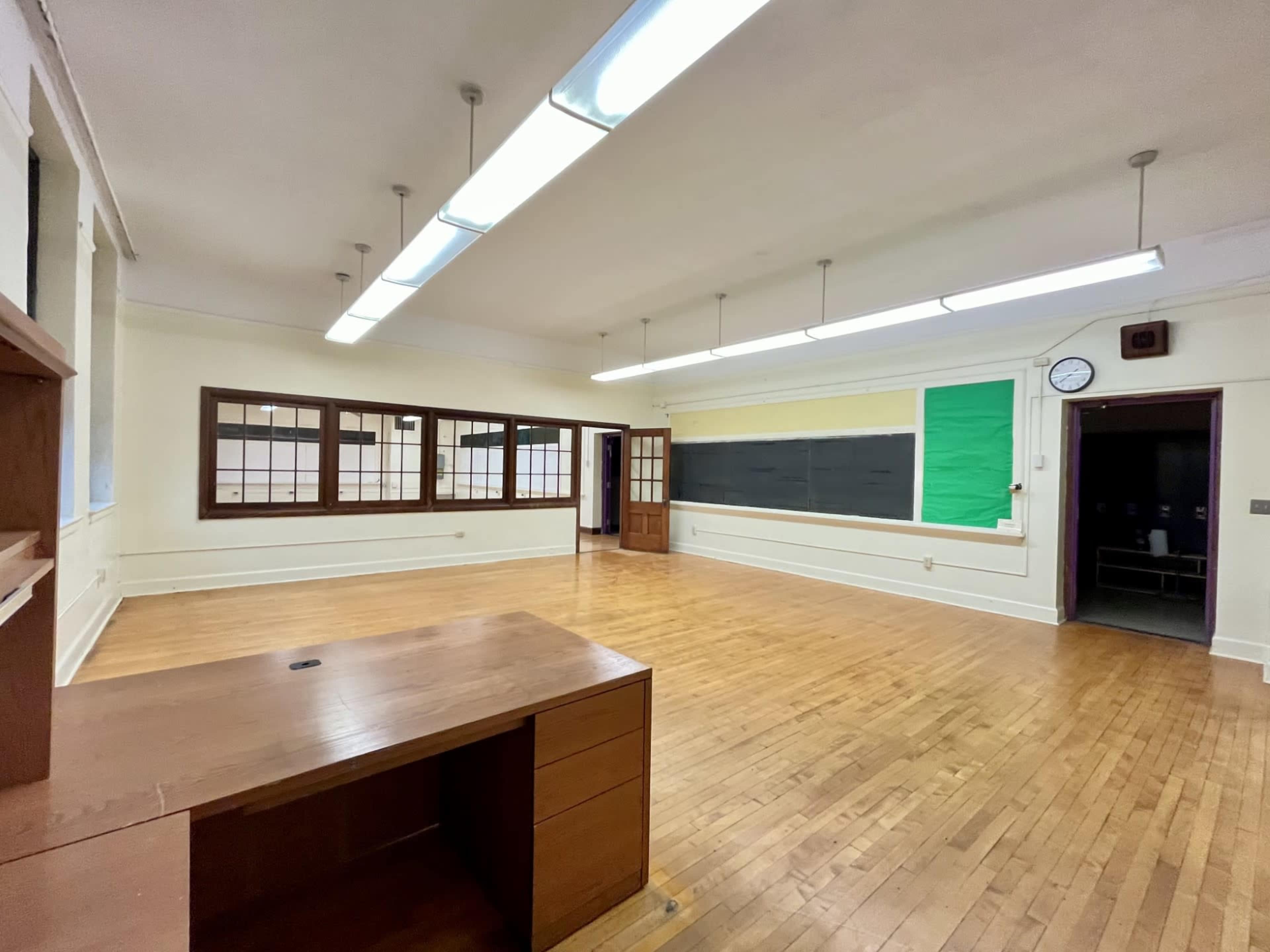 The image shows a spacious, empty classroom featuring wooden flooring, large windows, and a chalkboard at the front.
