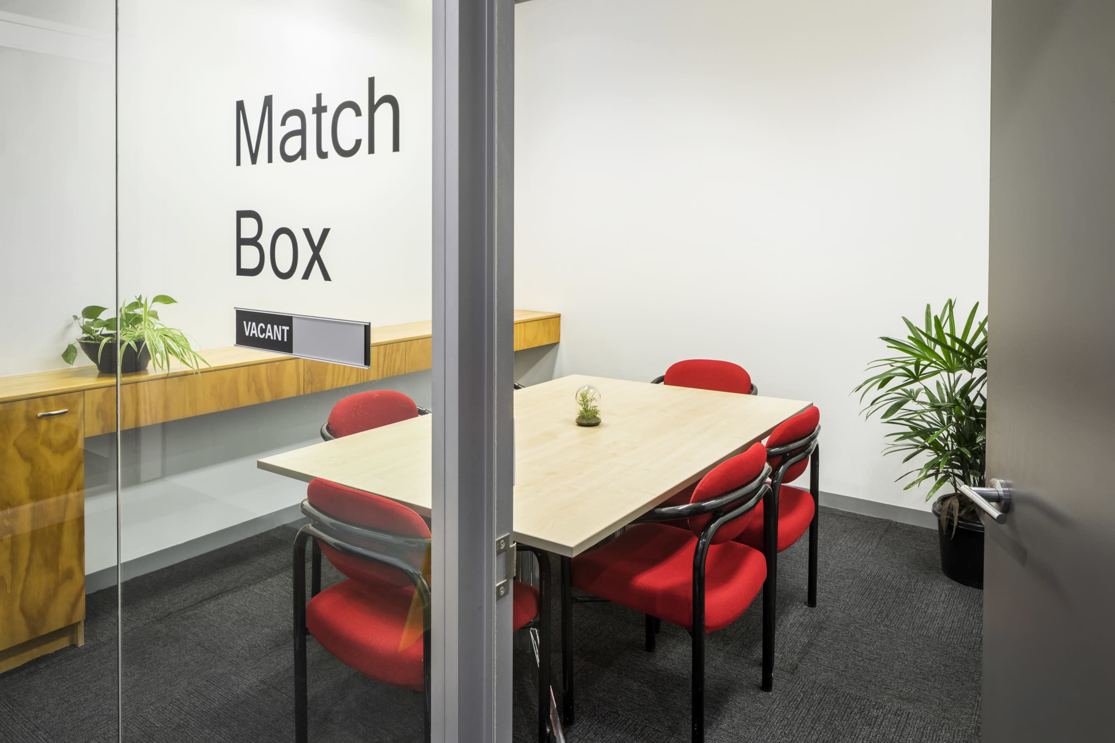 The image shows a vacant meeting room labeled "Match Box," featuring a rectangular table with four red chairs and a few indoor plants.