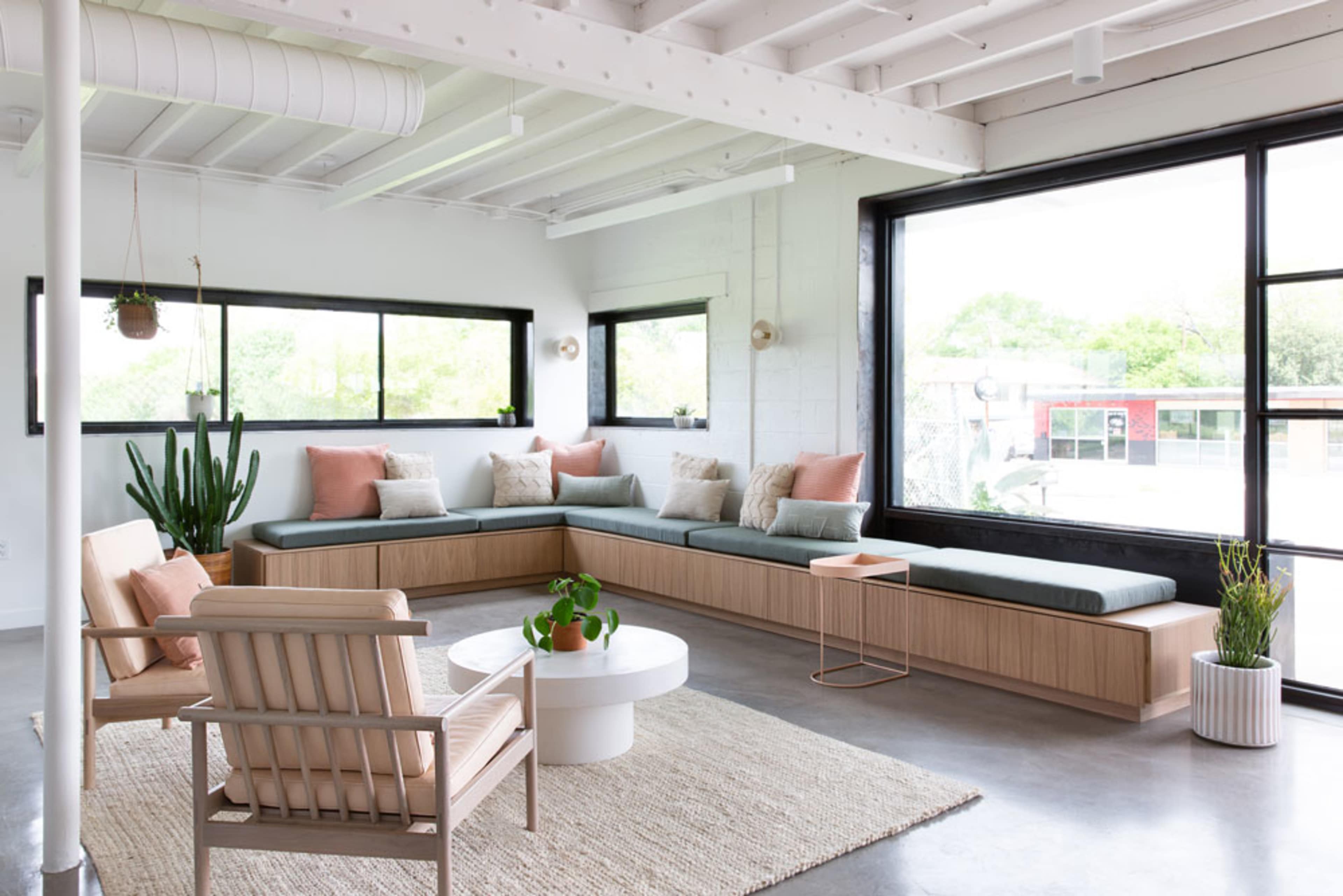 A bright and modern seating area features a large built-in bench with cushions, surrounded by windows and complemented by plants and a round coffee table.