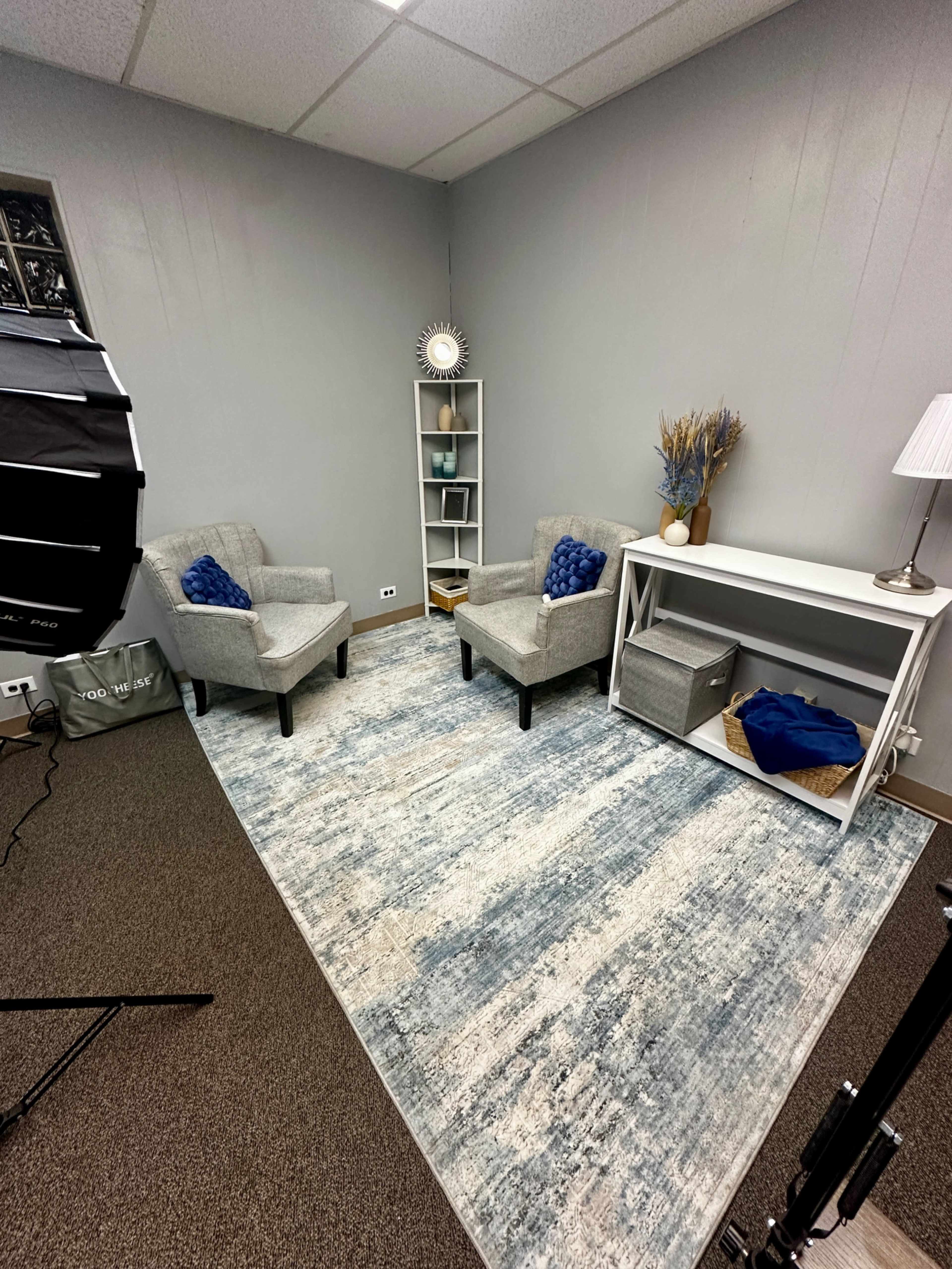 The image shows a small, neatly arranged room with two gray chairs facing each other, a white shelving unit, and a rug on the floor.